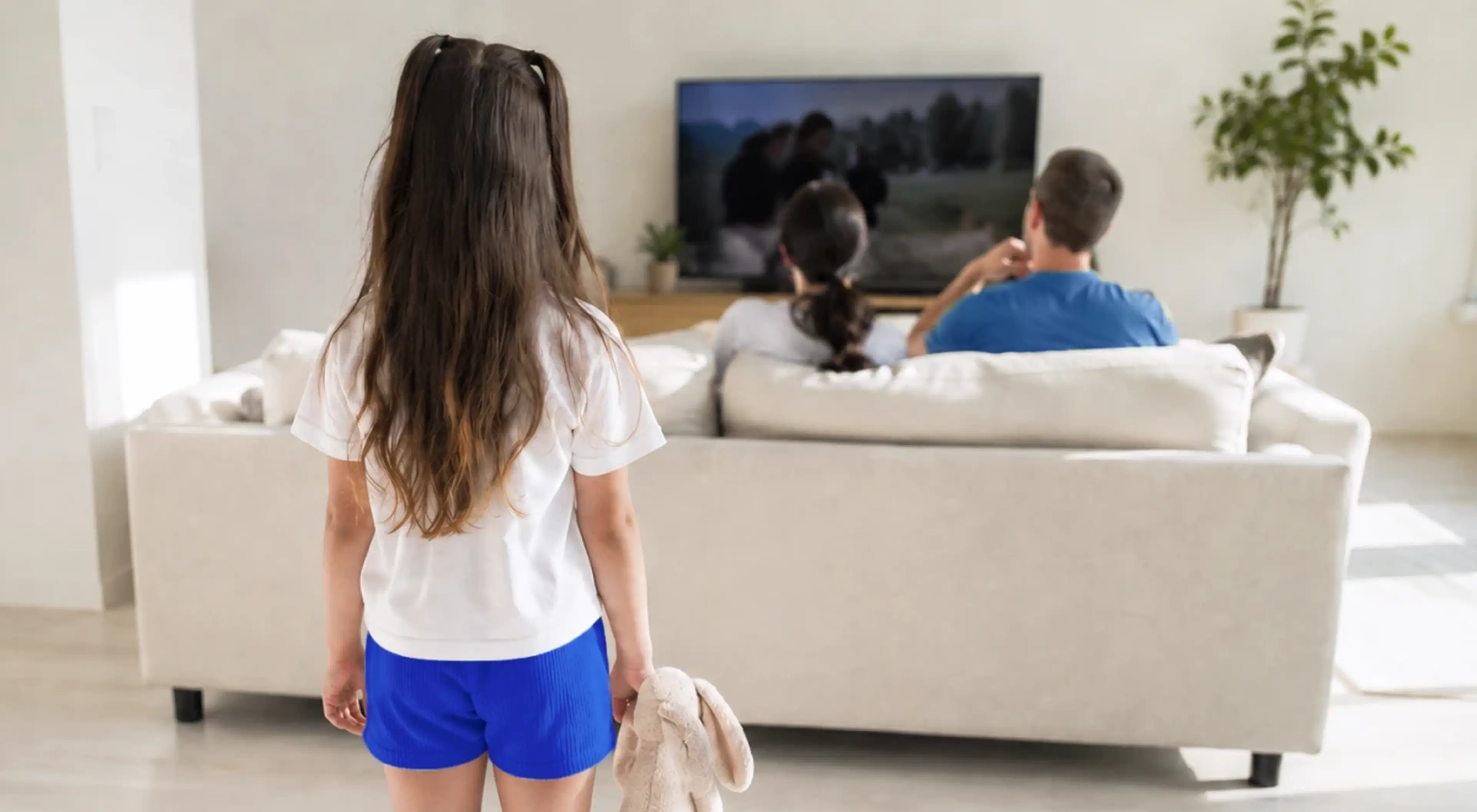 Young girl holding a stuffed bunny standing alone in a living room while her parents sit on a couch watching TV, illustrating child neglect and generational trauma