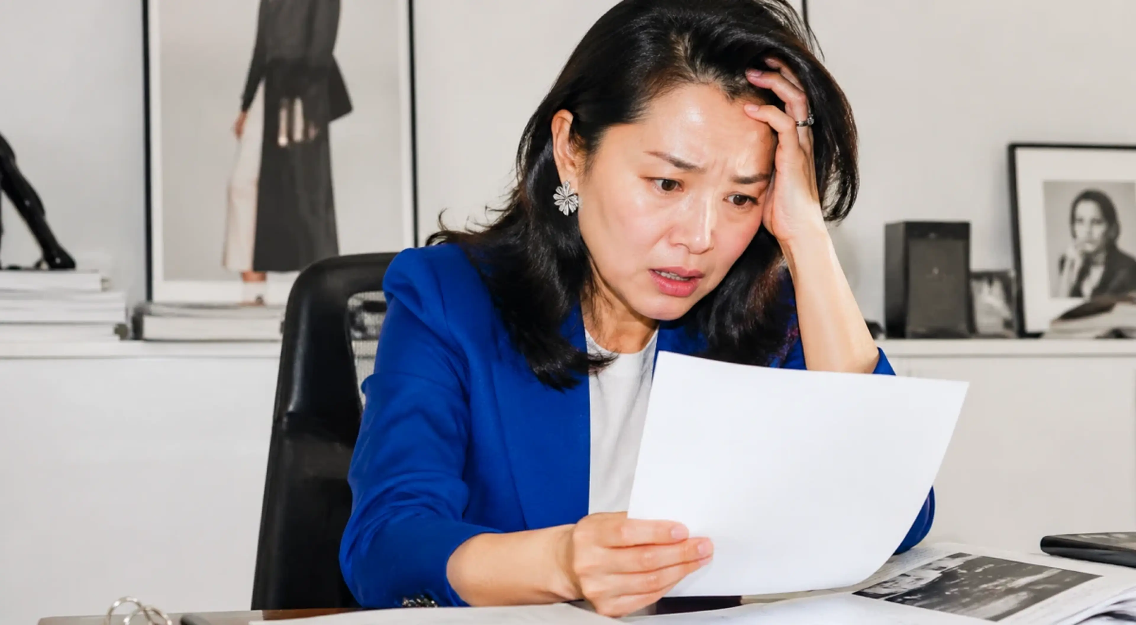 Stressed woman in a blue blazer sitting at a desk holding a document with a worried expression, hand on her forehead, with framed photos and fashion artwork on the wall behind her