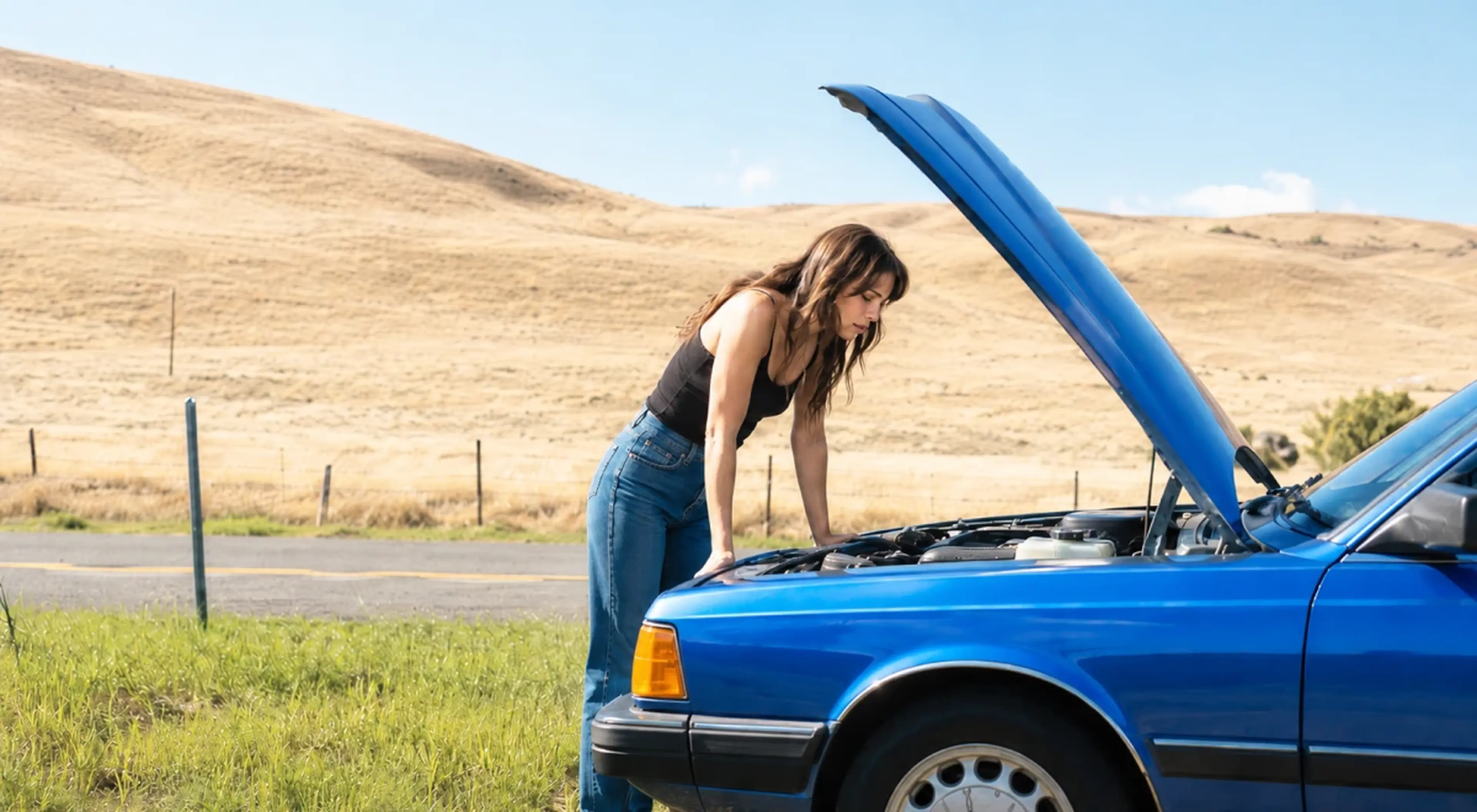 _Young woman leaning over the open hood of a broken down blue car on a rural roadside, representing external conflict and unexpected problems