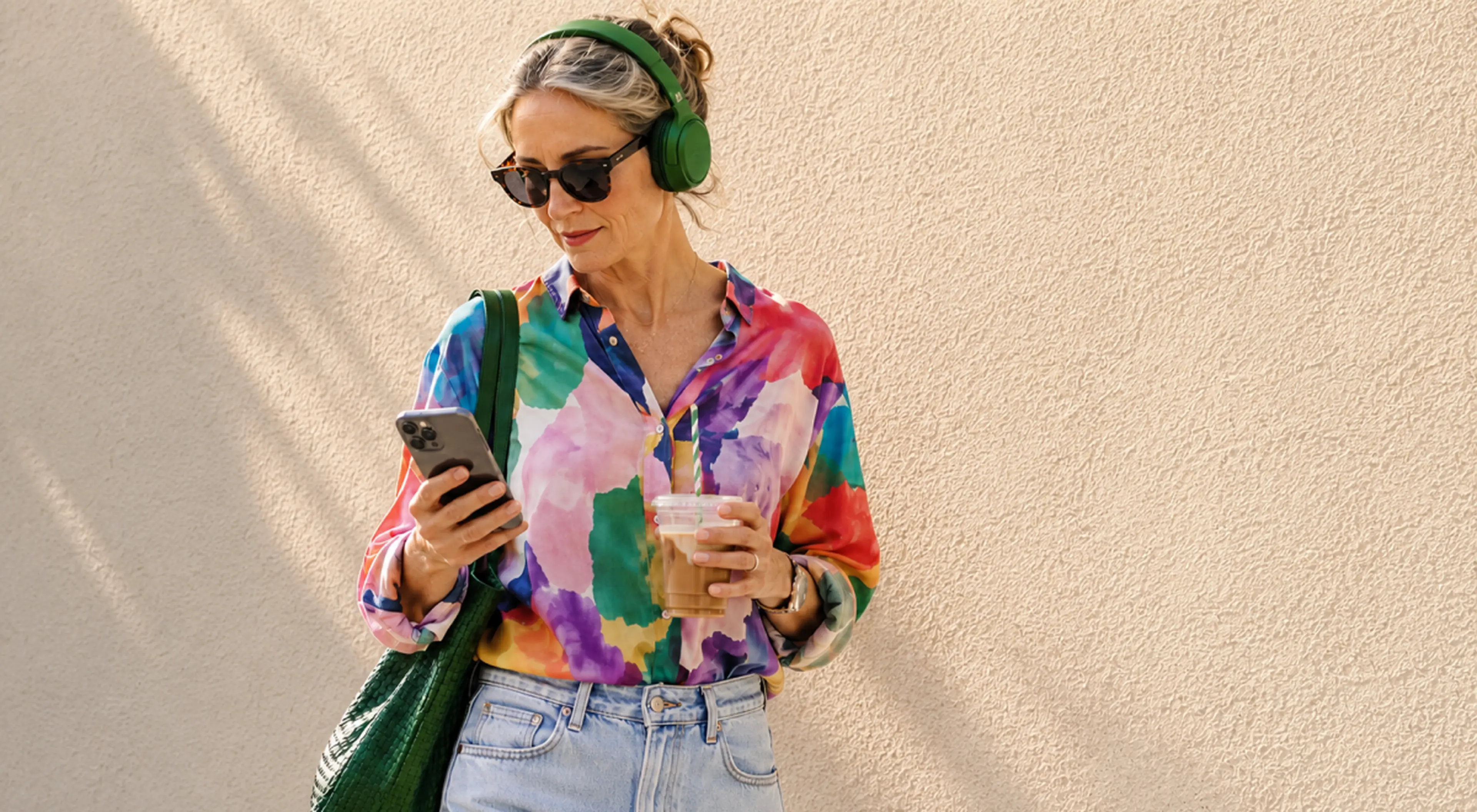 Woman in green headphones and a colorful blouse holding an iced coffee and smartphone against a beige wall, ready to listen to 20 books in 20 days