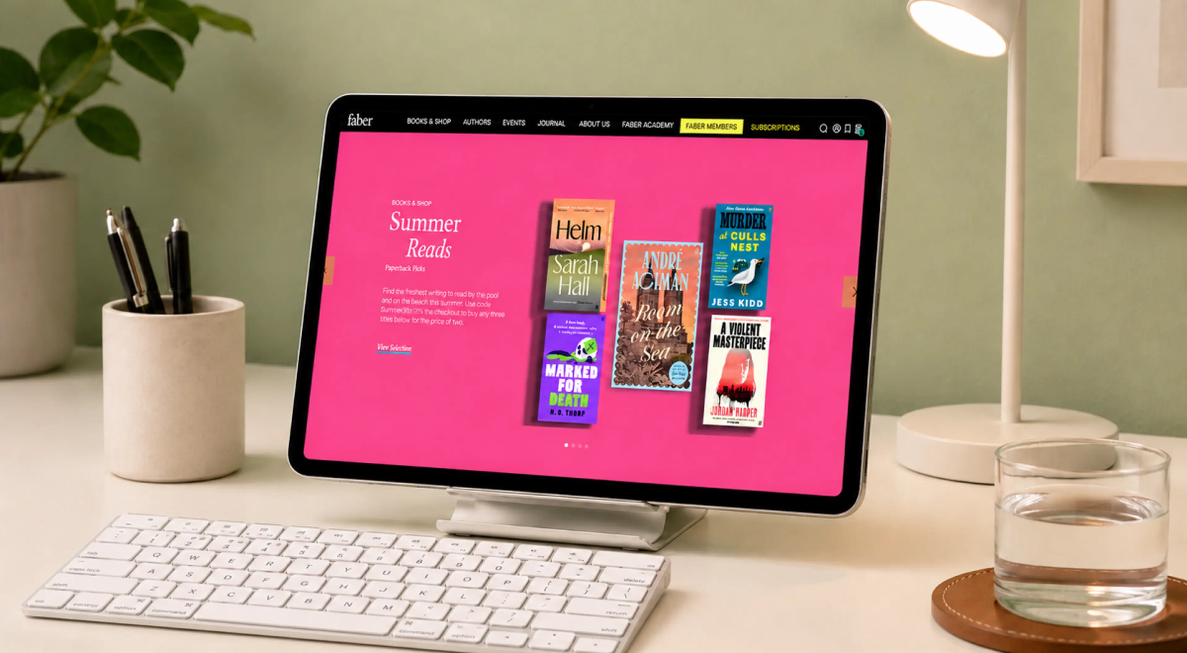 Faber and Faber website Summer Reads page shown on a tablet on a desk with keyboard, pen holder, glass of water and plant against a green wall