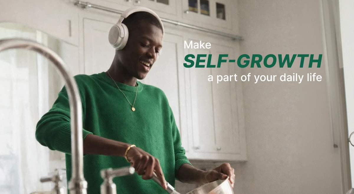 Smiling man in a green sweater wearing white headphones doing dishes in a bright kitchen while listening to one of the best audiobooks for daily self-growth