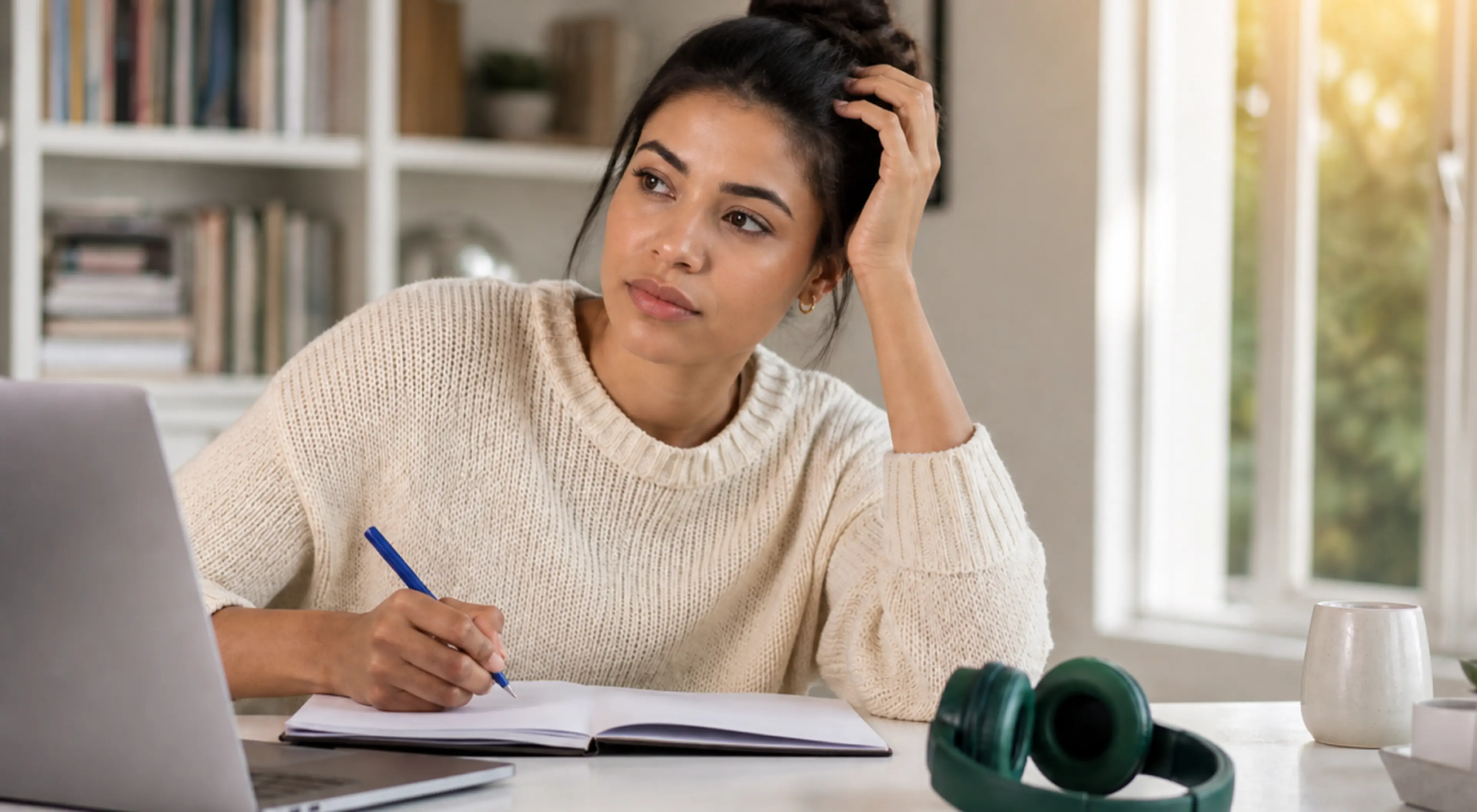 Thoughtful woman in a cream sweater writing in a notebook at a desk with a laptop, headphones and mug, pondering how to start writing a book by a sunny window