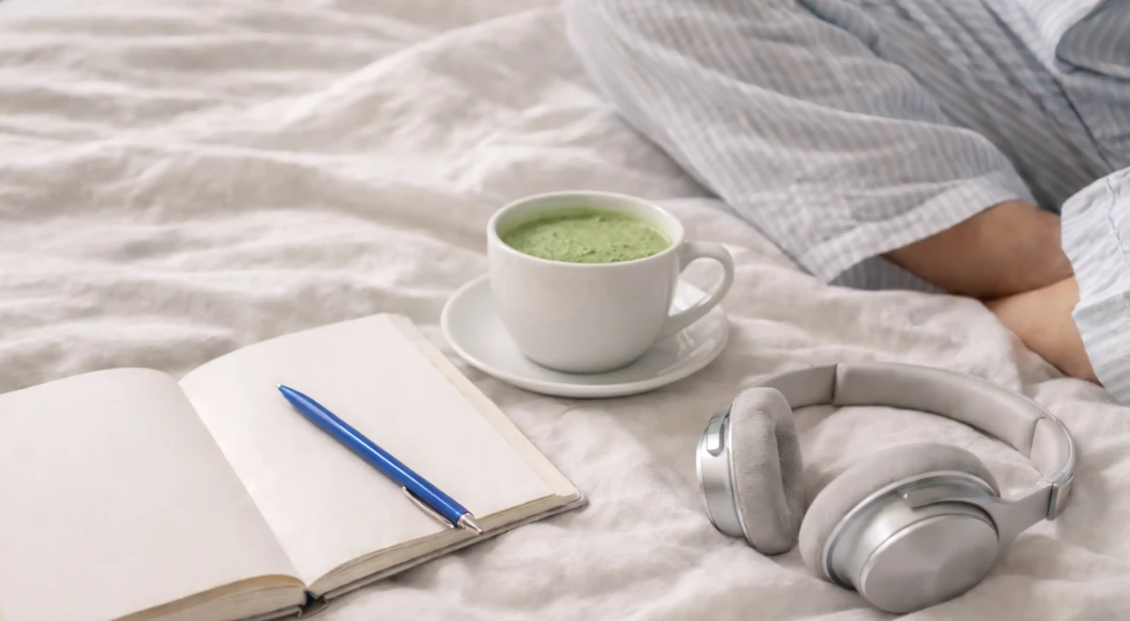 Person in bed with open notebook, blue pen, cup of green matcha, and silver headphones on white linen as part of a successful person's morning routine