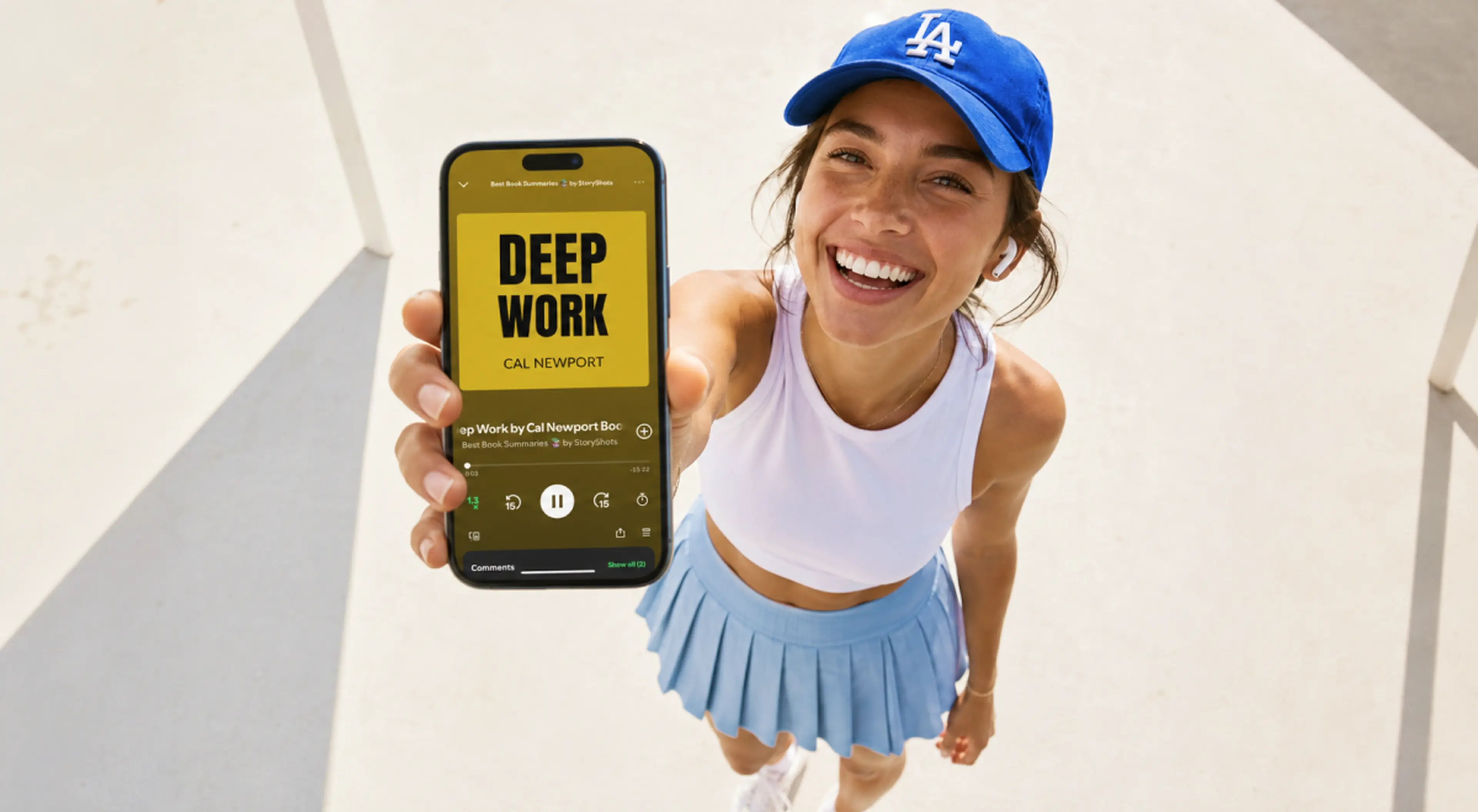 Smiling woman in a blue LA cap and white top holds up her phone showing the Deep Work by Cal Newport audiobook on the StoryShots app while listening outdoors