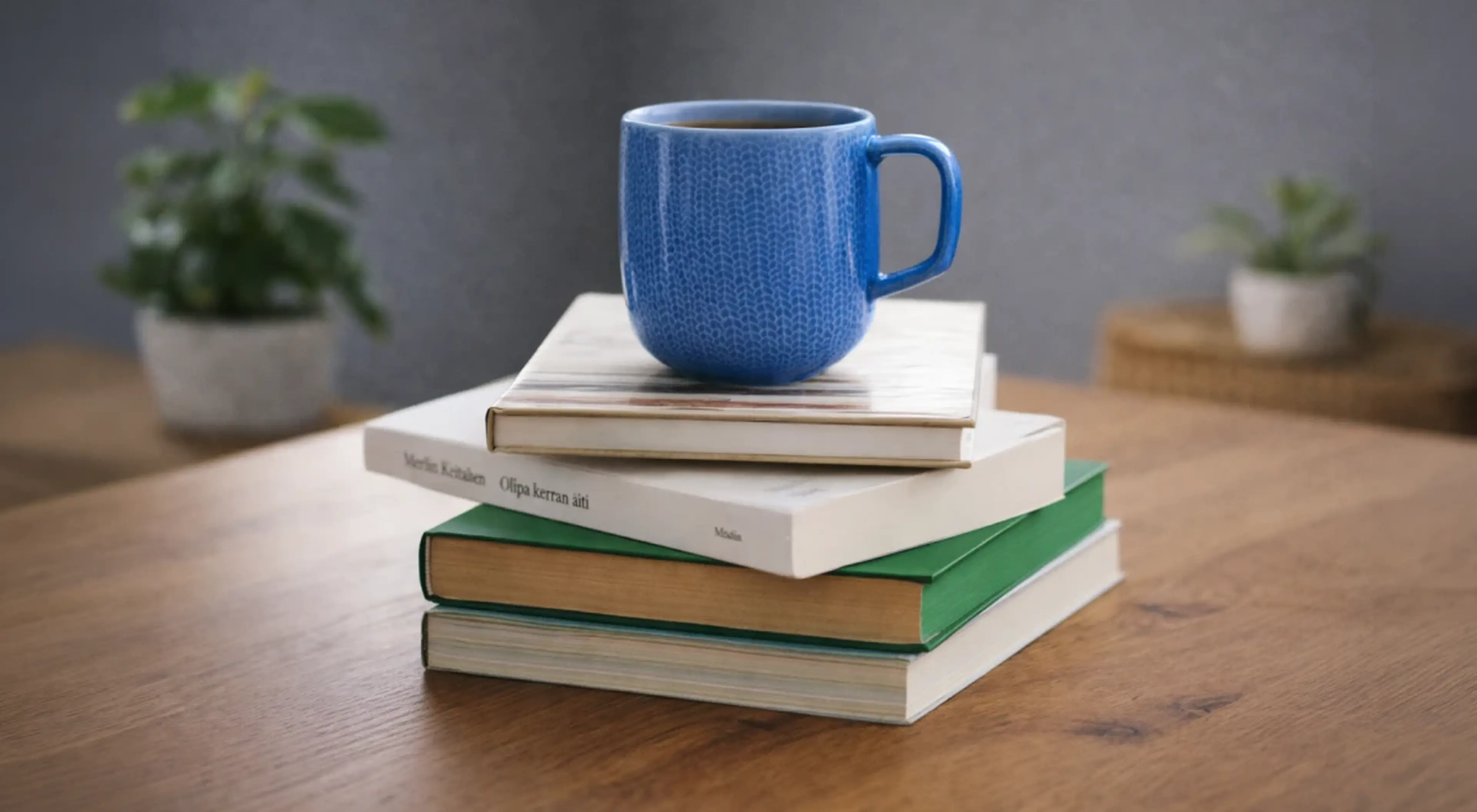 Blue ceramic mug resting on a stack of books on a wooden table, with potted plants and a gray background — a cozy reading habit setup