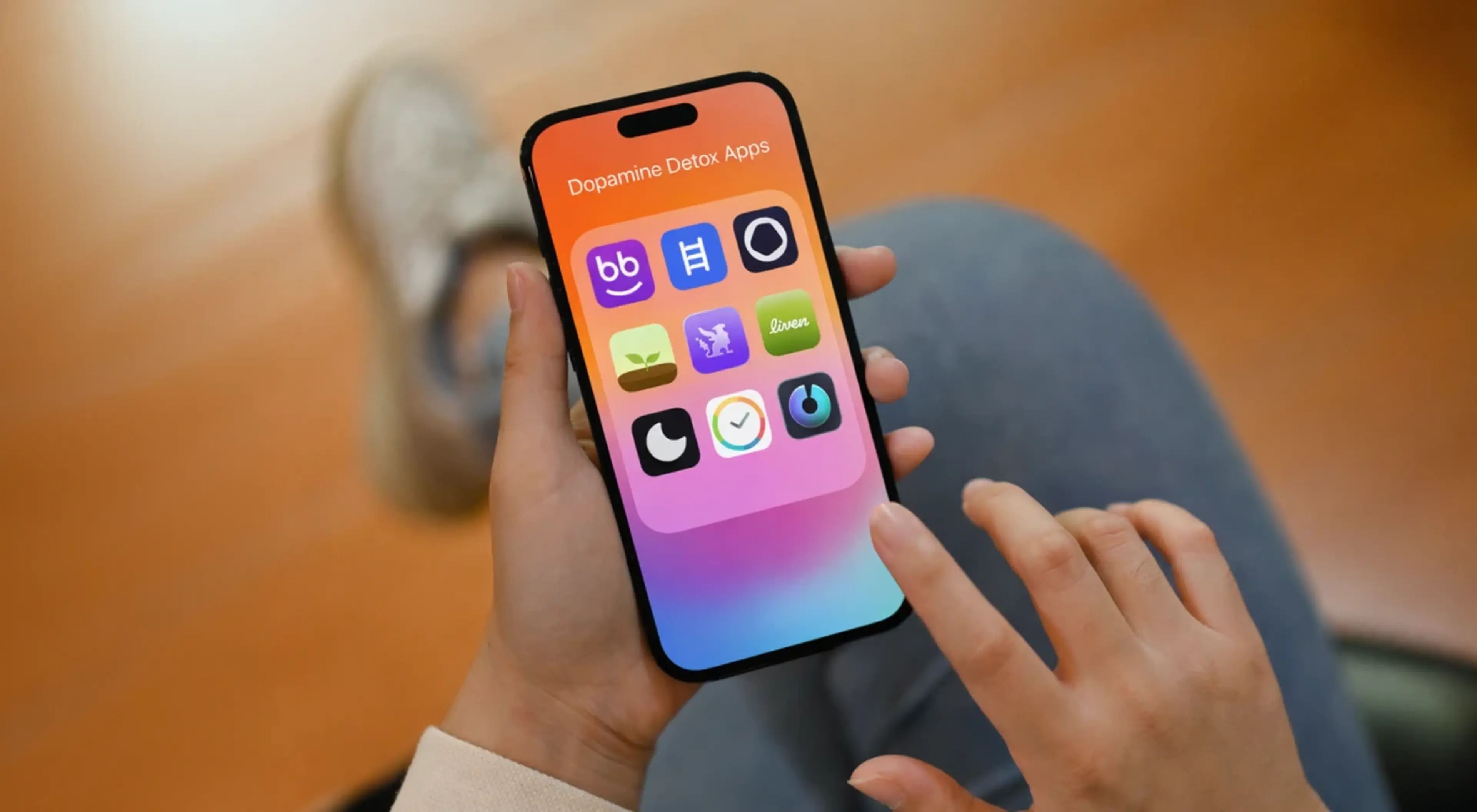 Person holding a smartphone displaying a dopamine detox apps folder with colorful app icons, sitting on a wooden floor with jeans visible in background
