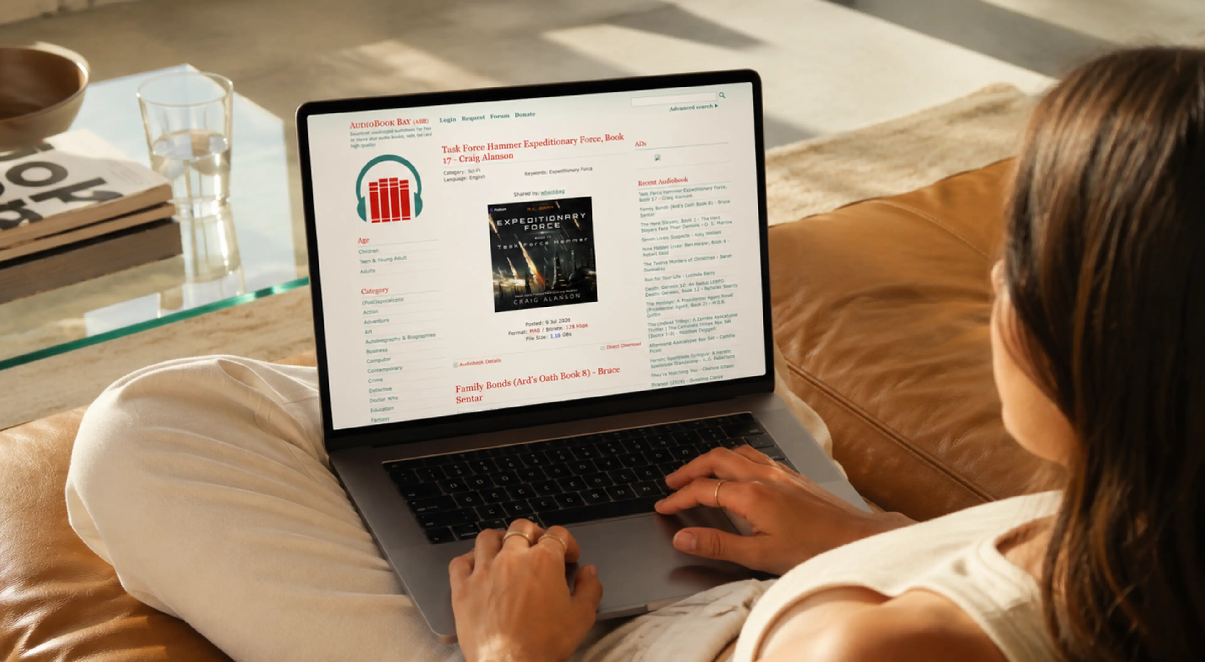 Woman relaxing on a tan leather couch browsing the AudioBookBay website on a laptop, viewing the Expeditionary Force audiobook by Craig Alanson beside a glass table with water and magazines