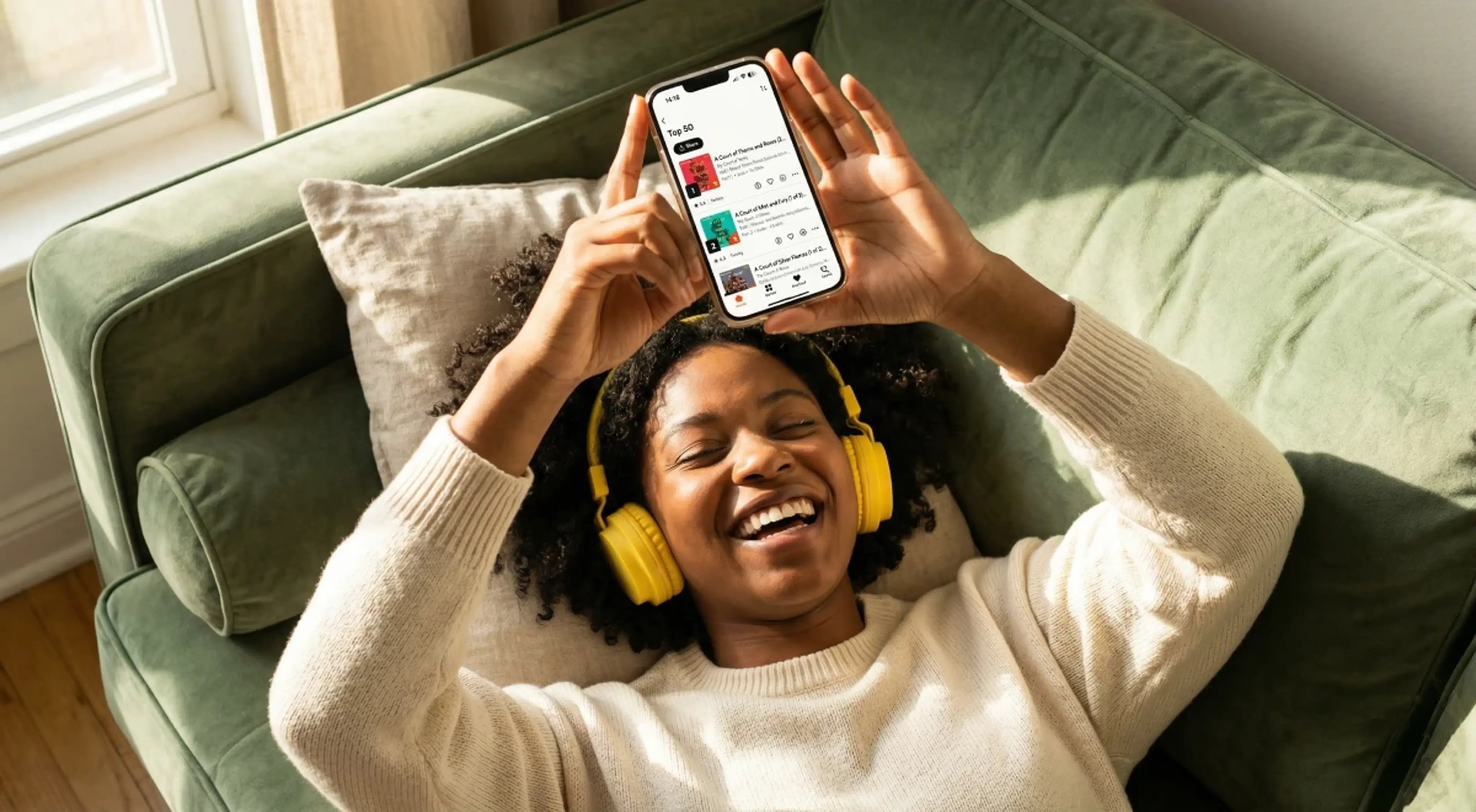 Smiling woman with yellow headphones lying on a green couch, holding up a smartphone with the Storytel audiobooks app, listening to an audiobook