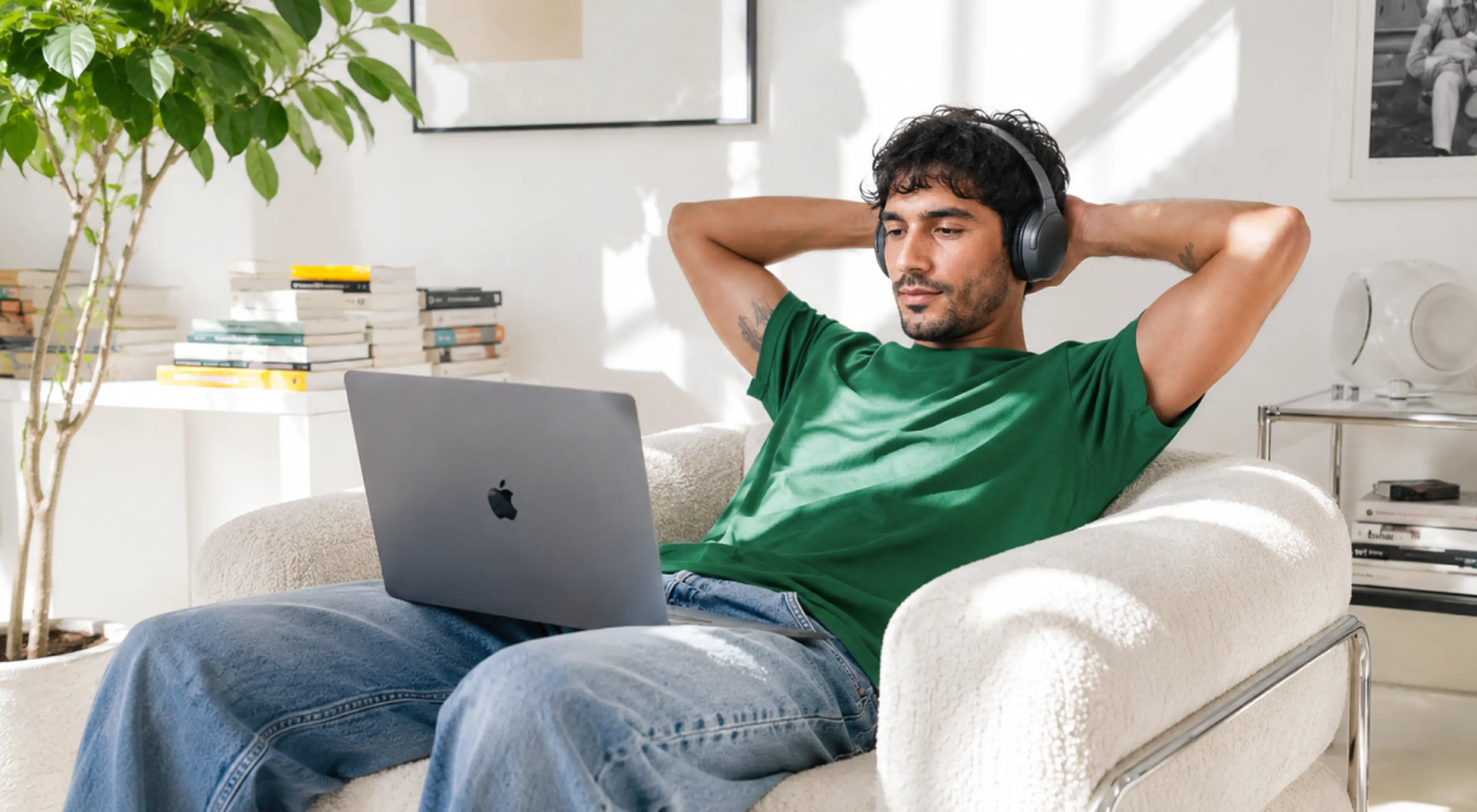 Man in green shirt and headphones relaxing on a white armchair with an Apple laptop, listening to self-help podcasts in a bright living room with books and a plant
