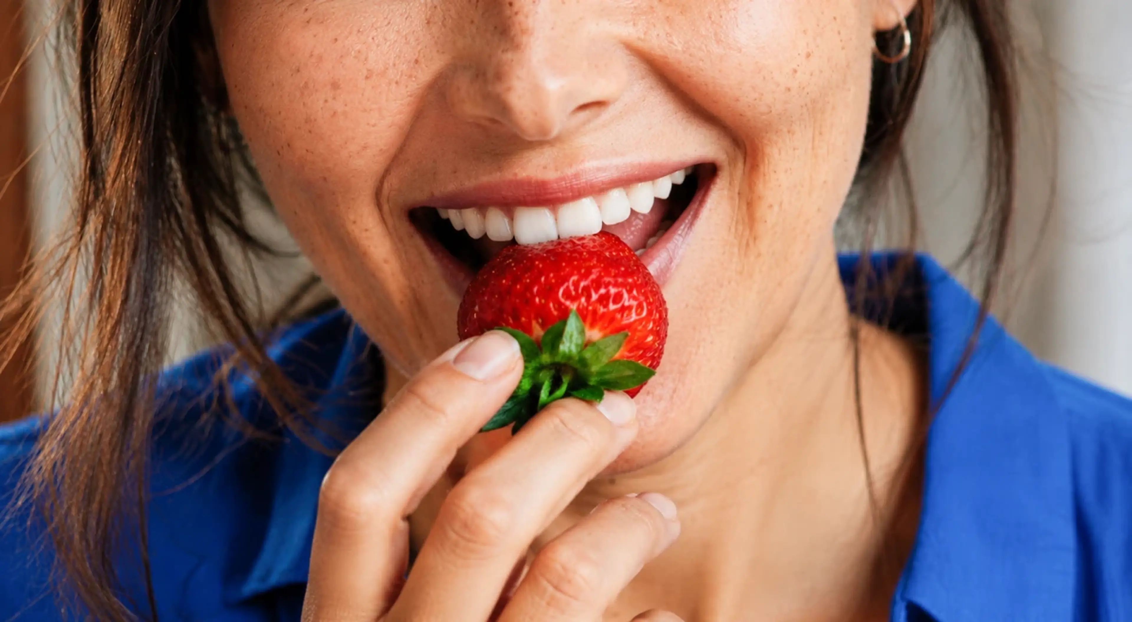 Woman in a blue shirt smiling while eating a fresh red strawberry, representing healthy eating and mindful nutrition habits
