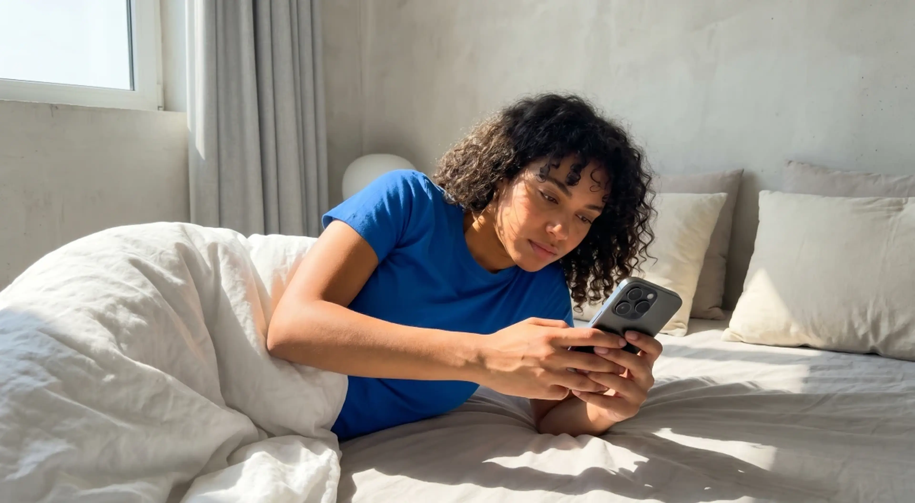 Young woman with curly hair in a blue t-shirt lying in bed scrolling on her smartphone in a bright bedroom, illustrating excessive phone use