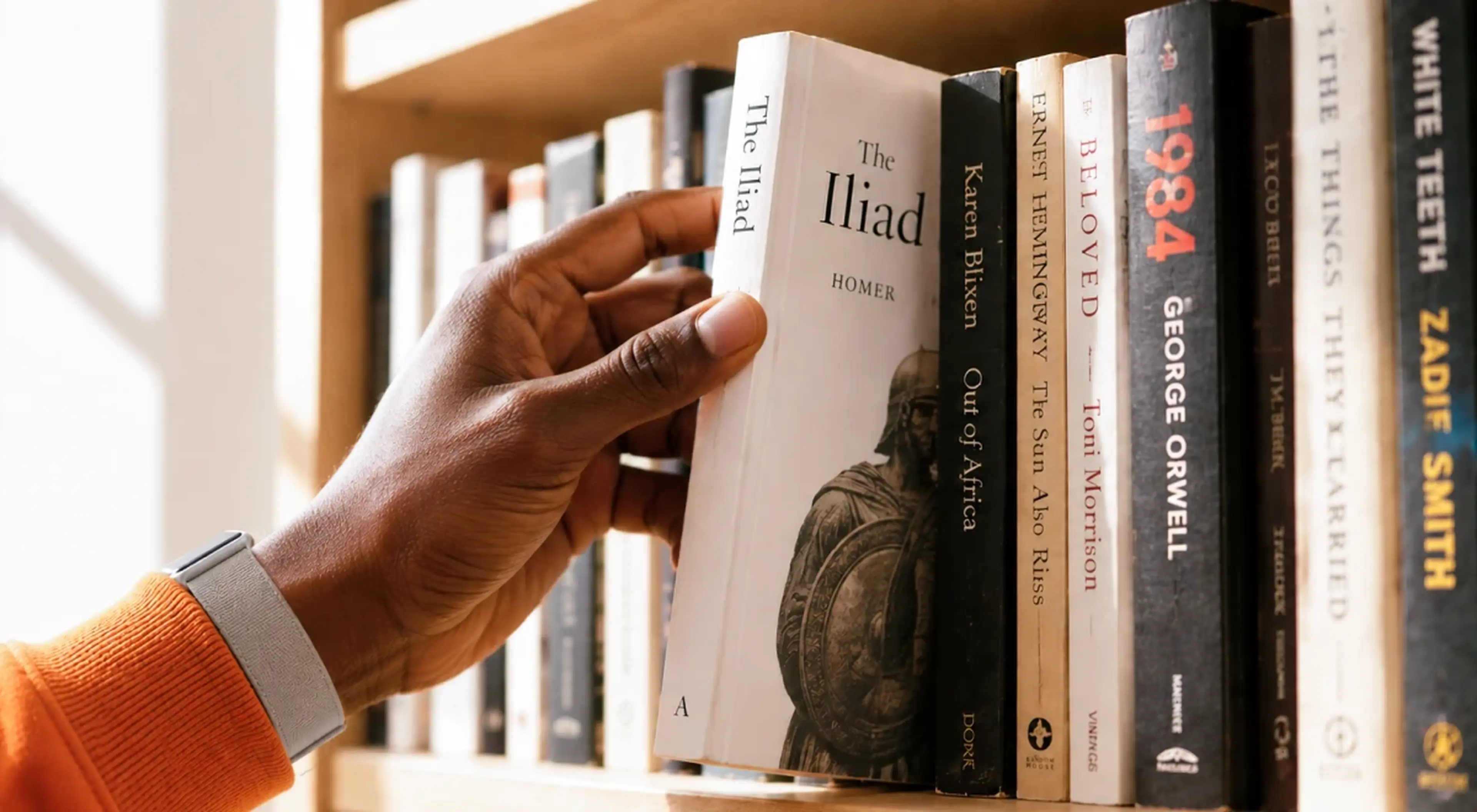 A hand in an orange sleeve pulling The Iliad by Homer from a wooden bookshelf filled with classic novels including 1984 by George Orwell and Beloved by Toni Morrison