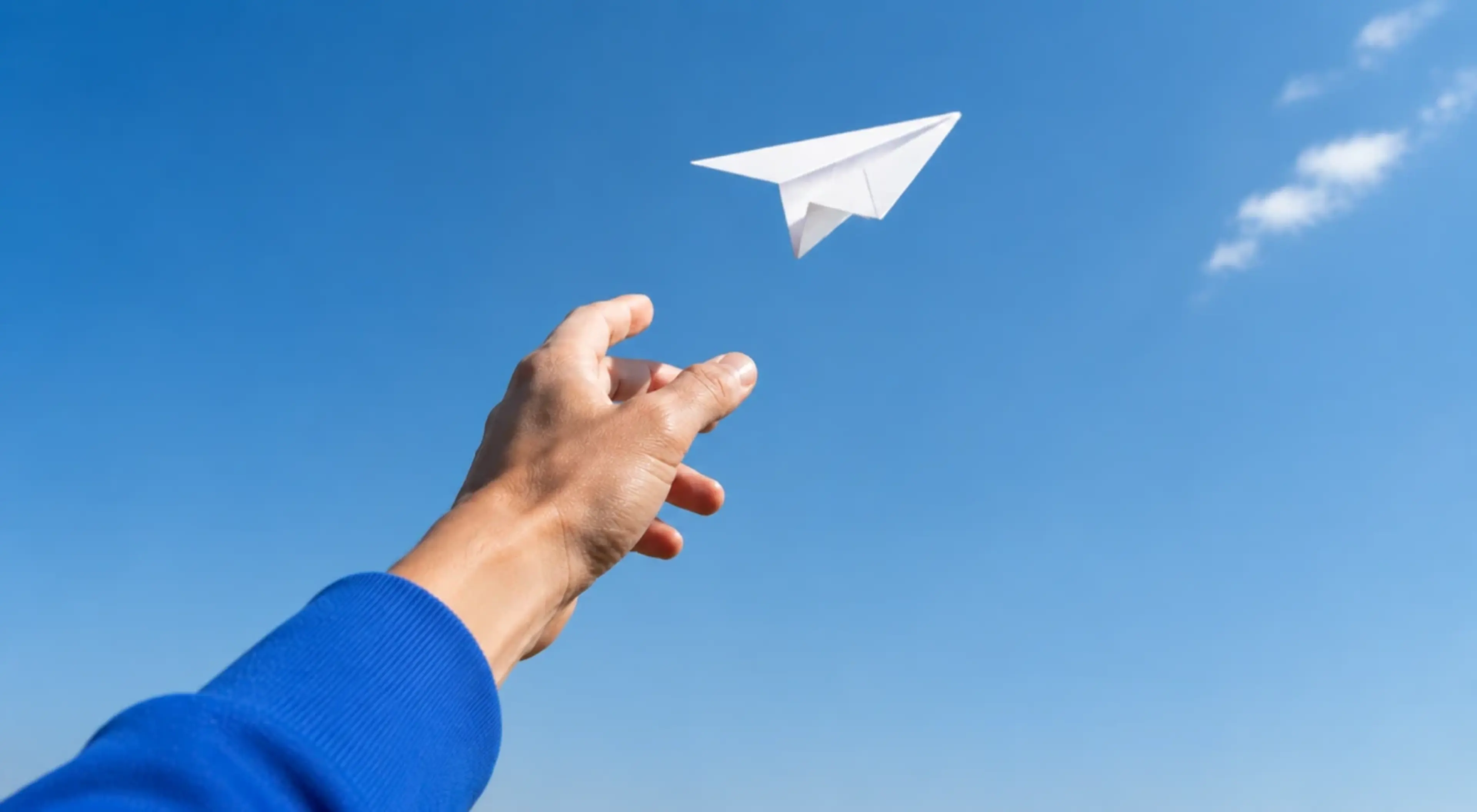 Hand in a blue sleeve releasing a paper plane into a clear blue sky, symbolizing the act of forgiving yourself and letting go of the past