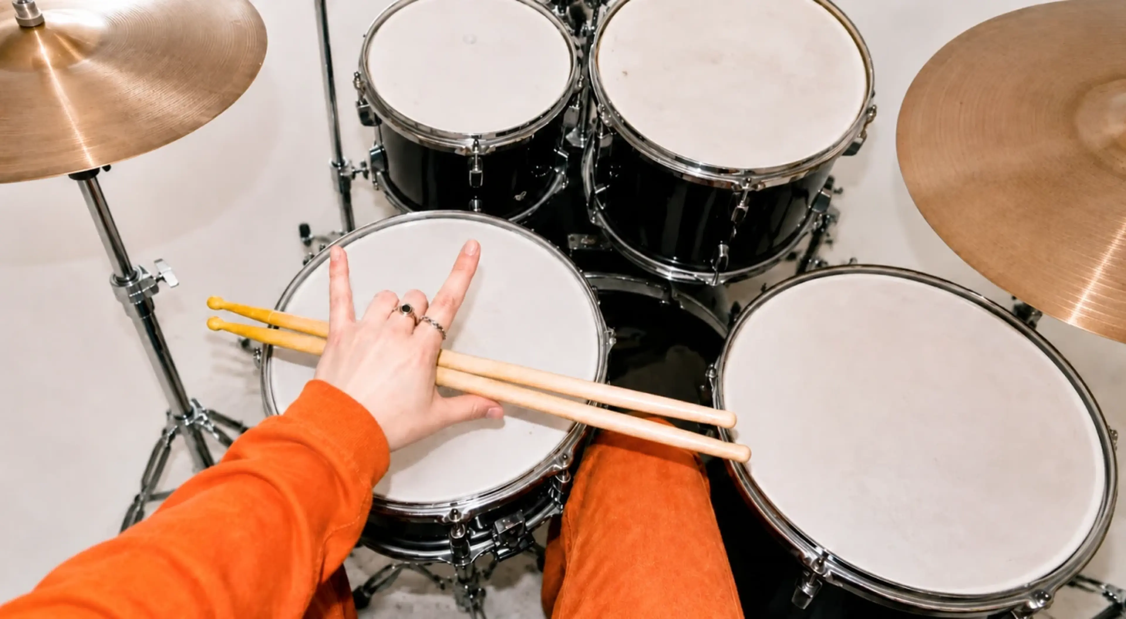 Drummer in an orange sleeve making a rock hand sign over a snare drum, holding wooden drumsticks, with a black drum kit and cymbals in the background