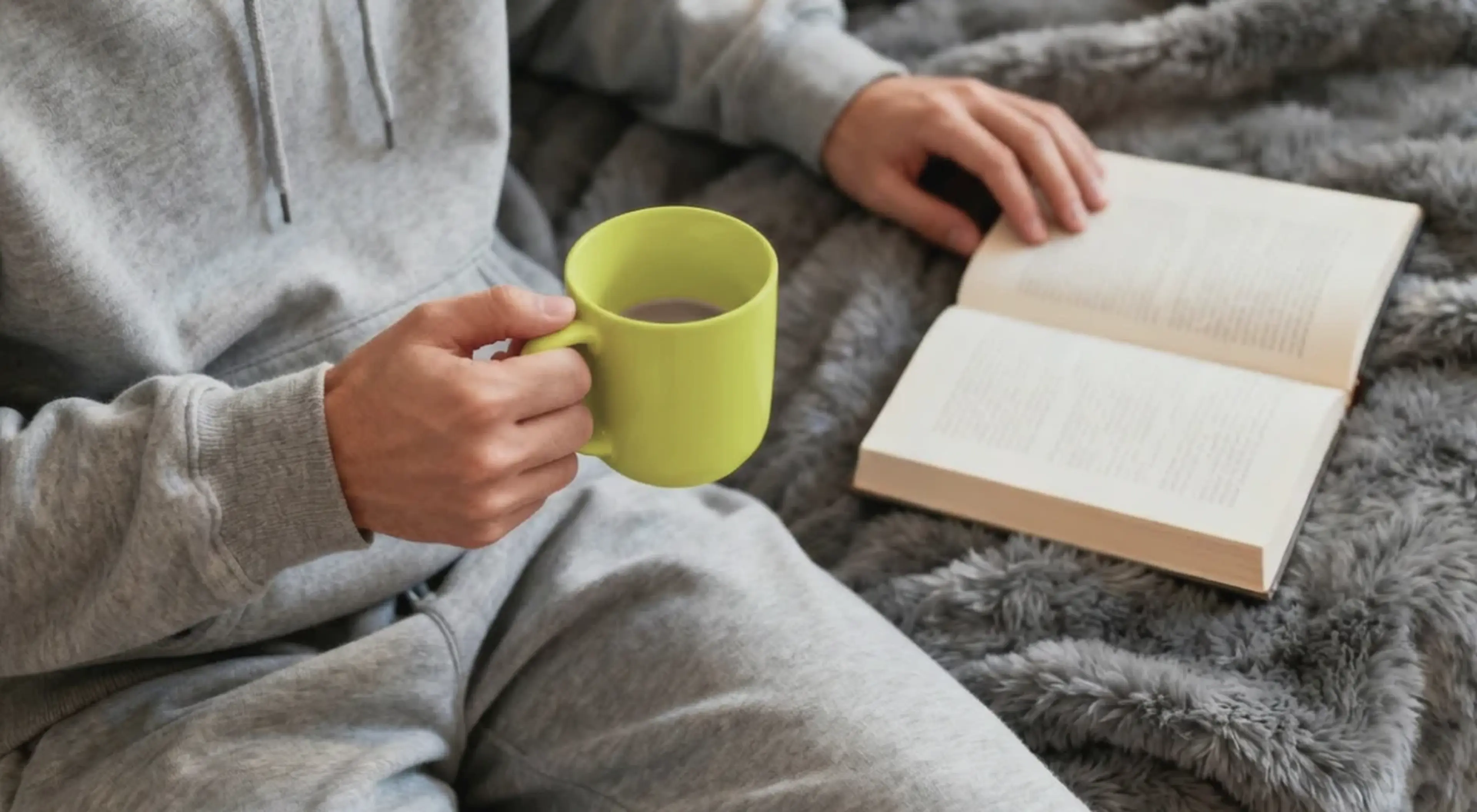 Person in gray hoodie holding a green mug and reading an open book on a fluffy gray blanket, illustrating a cozy daily reading habit