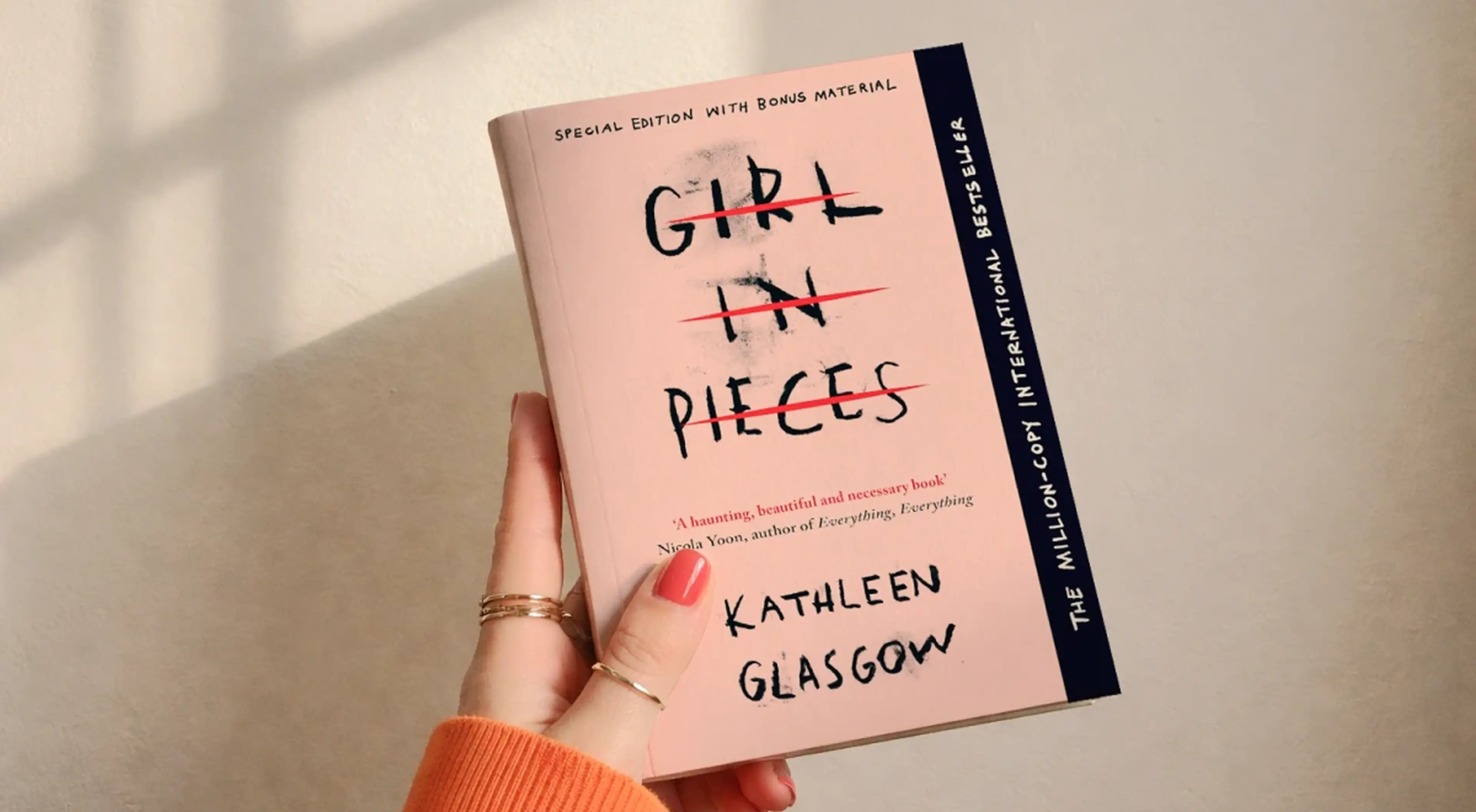 _Hand with red nails and gold rings holding Girl in Pieces by Kathleen Glasgow against a sunlit beige wall — special edition book review pick