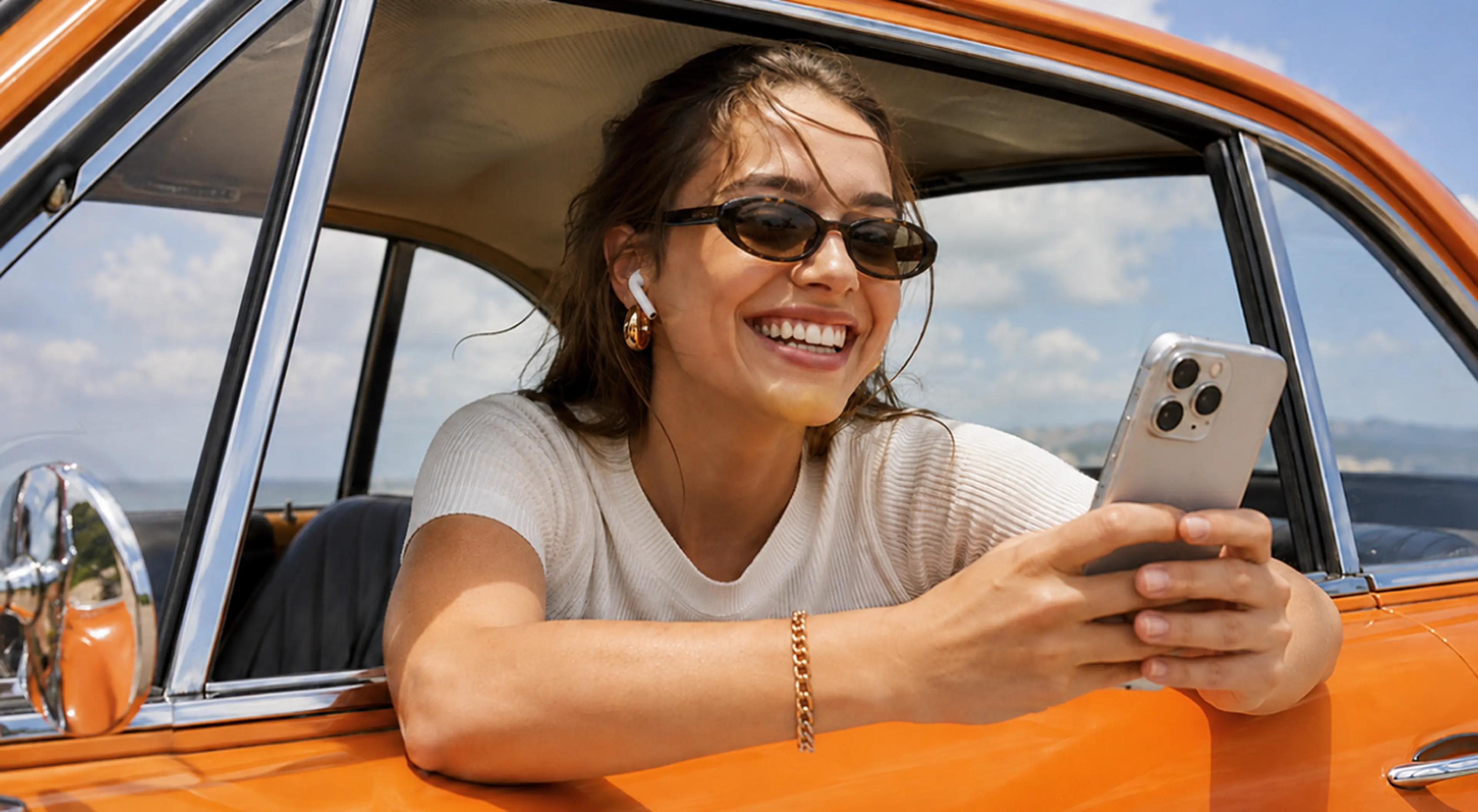 Smiling woman in sunglasses and AirPods leaning out of an orange vintage car window using an iPhone with a microlearning app under a bright blue sky
