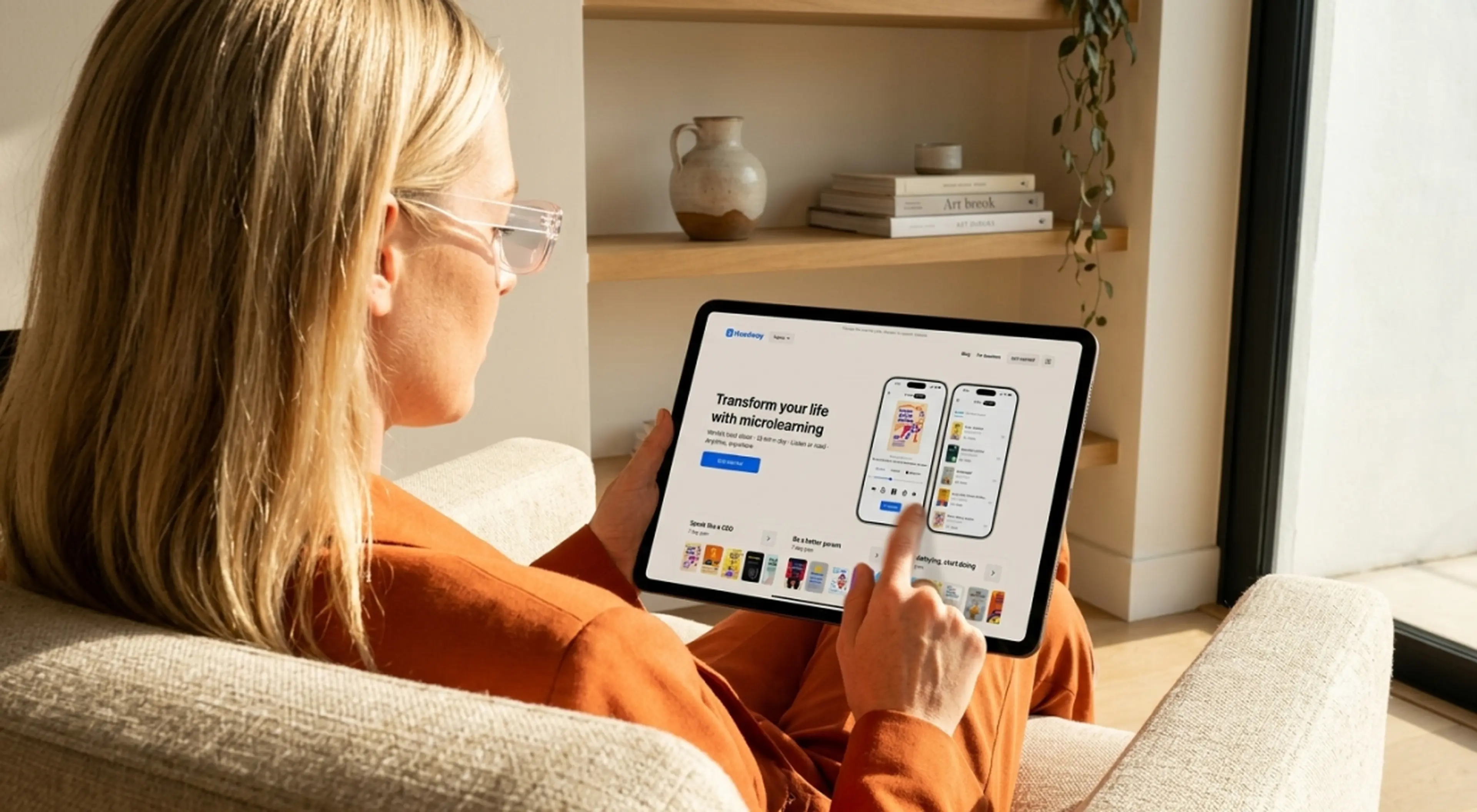 Blonde woman in an orange sweater and glasses browsing the Headway microlearning app on a tablet while relaxing on a beige couch by a bookshelf, reflecting microlearning trends