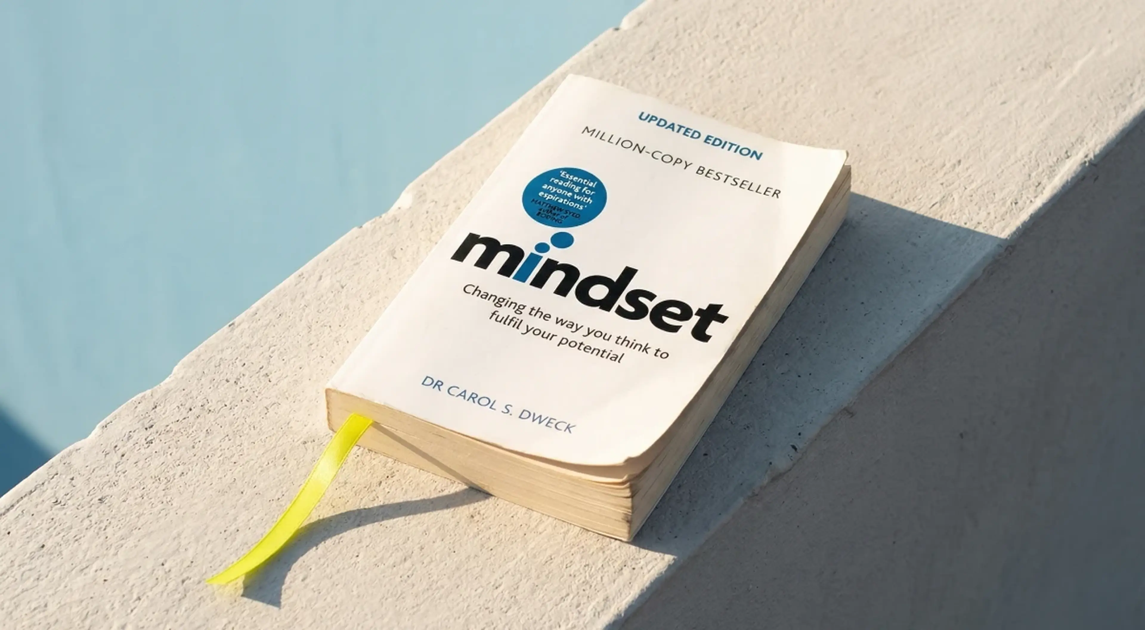 Mindset by Dr Carol SDweck growth mindset book with yellow bookmark resting on a sunlit concrete ledge against a light blue background