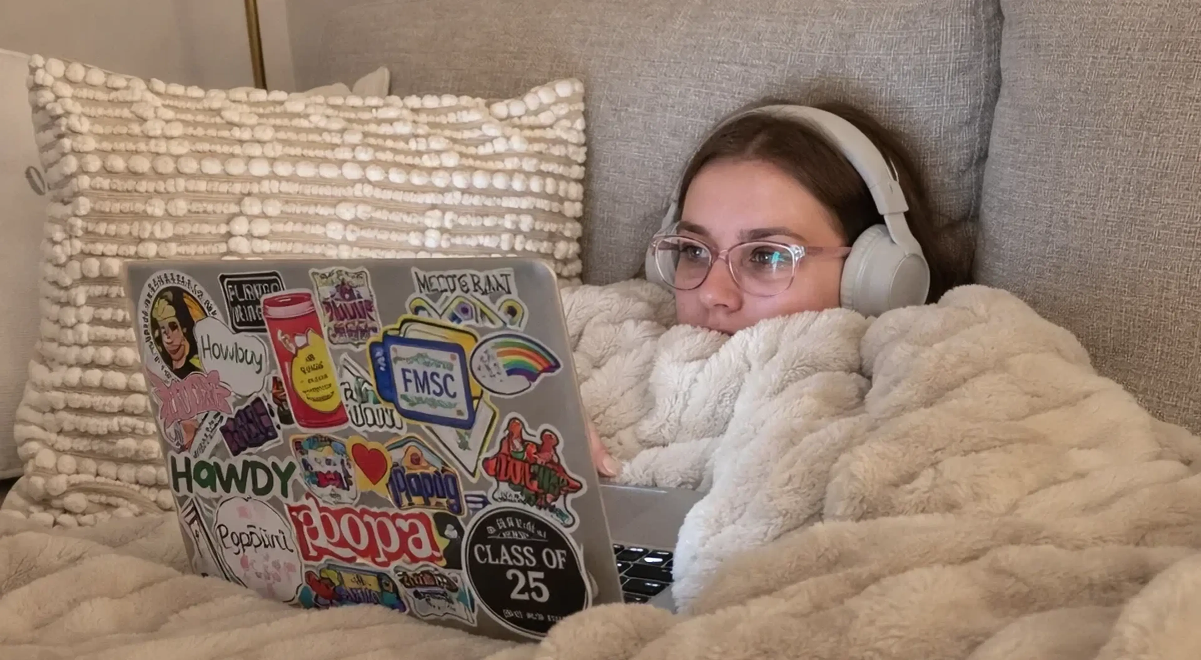 Young woman with headphones and glasses lying on a couch under a fluffy blanket, using a sticker-covered laptop — reflecting cozy digital habits and social media use