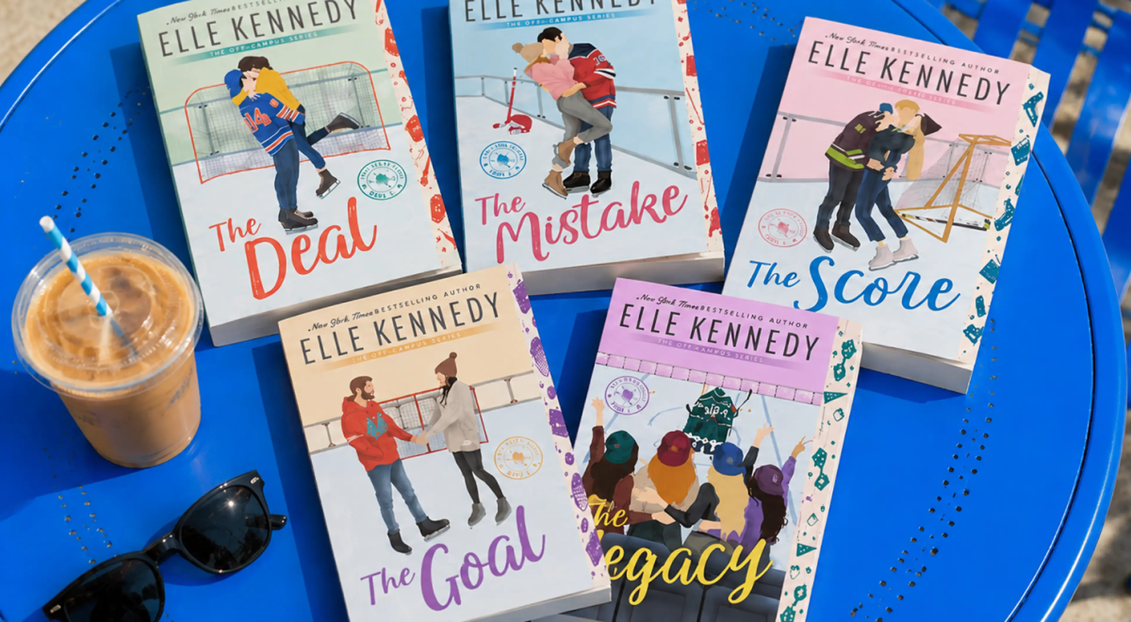 Elle Kennedy's Off-Campus book series paperbacks including The Deal, The Mistake, The Score, The Goal and The Legacy arranged on a blue table beside an iced coffee and sunglasses