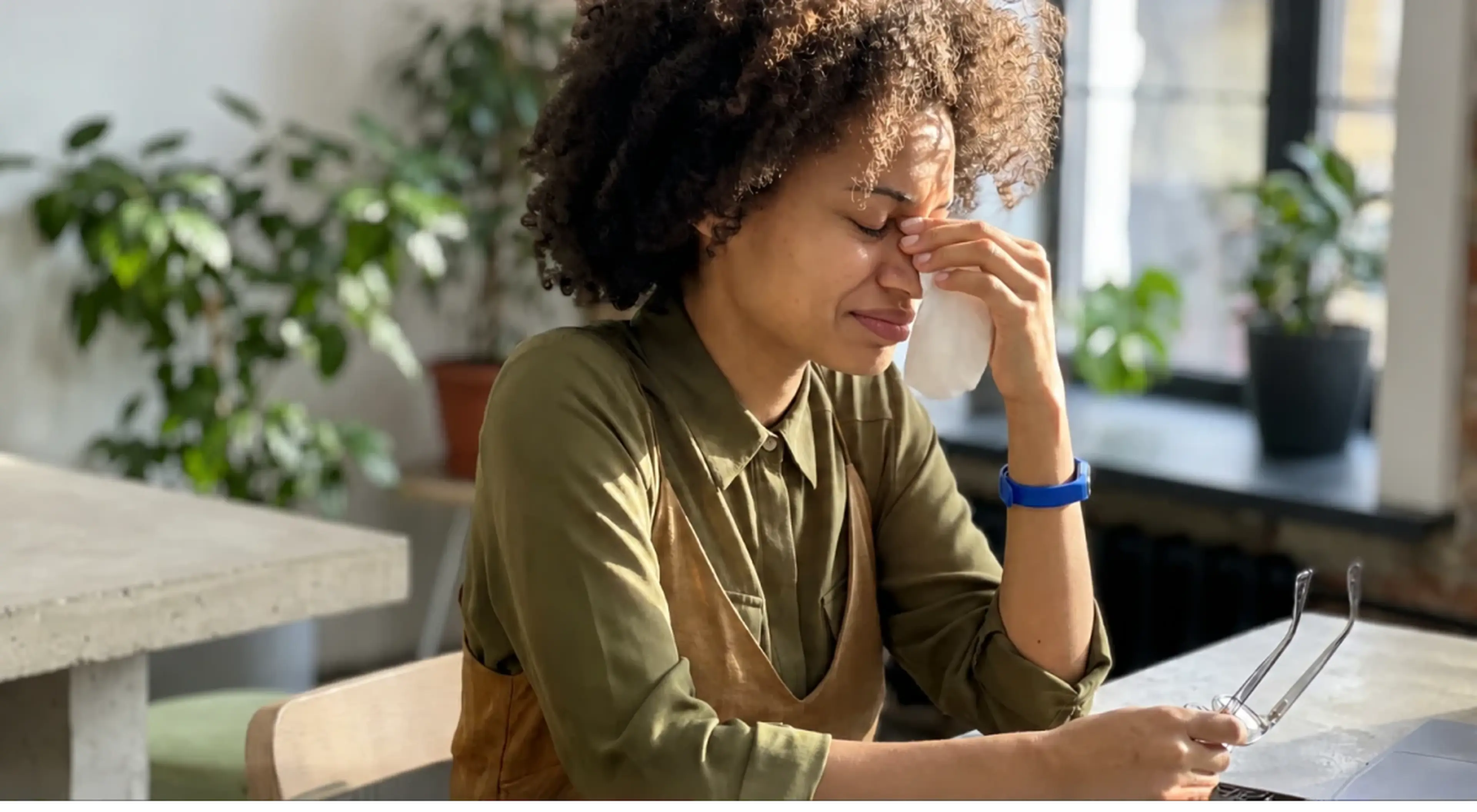 A woman with curly hair sitting at a desk, pressing her fingers to her nose with eyes closed, appearing stressed or overwhelmed, with green plants and a window in the background