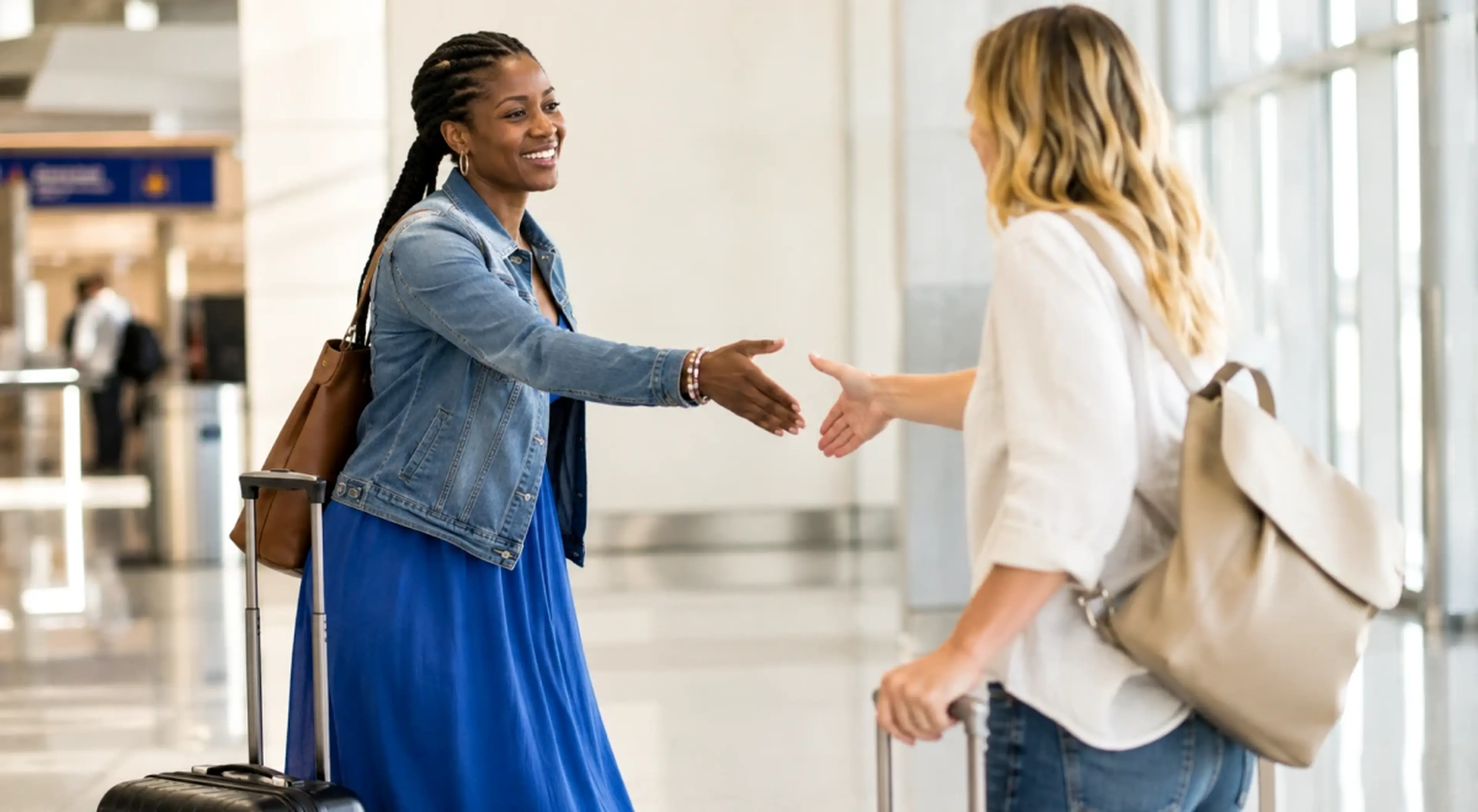 Two women with luggage reaching out to shake hands and greet each other warmly in an airport terminal, showing how to talk to anyone