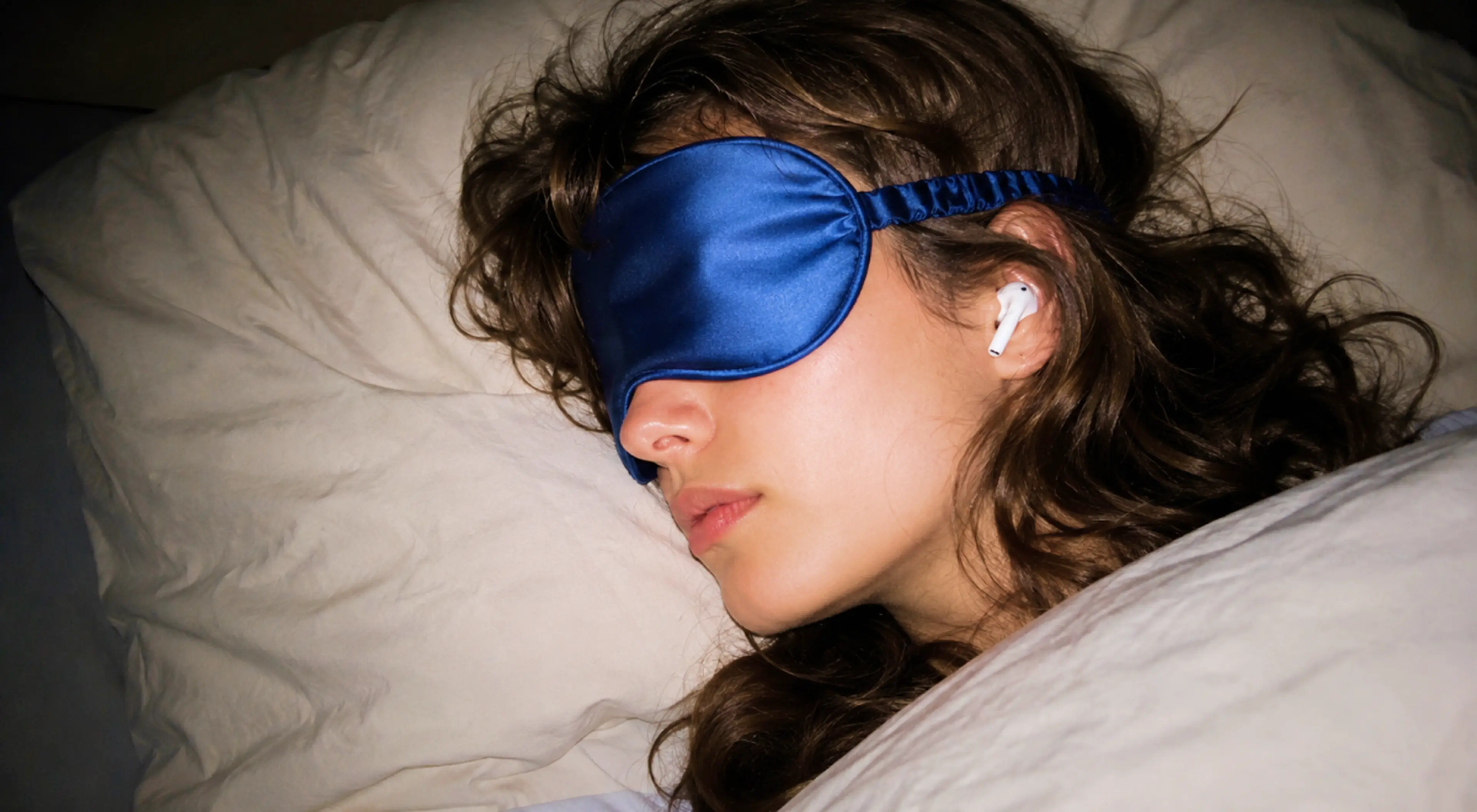 Woman with curly brown hair resting in bed wearing a blue satin sleep mask and a wireless earbud, listening to sleep stories for adults on white pillows