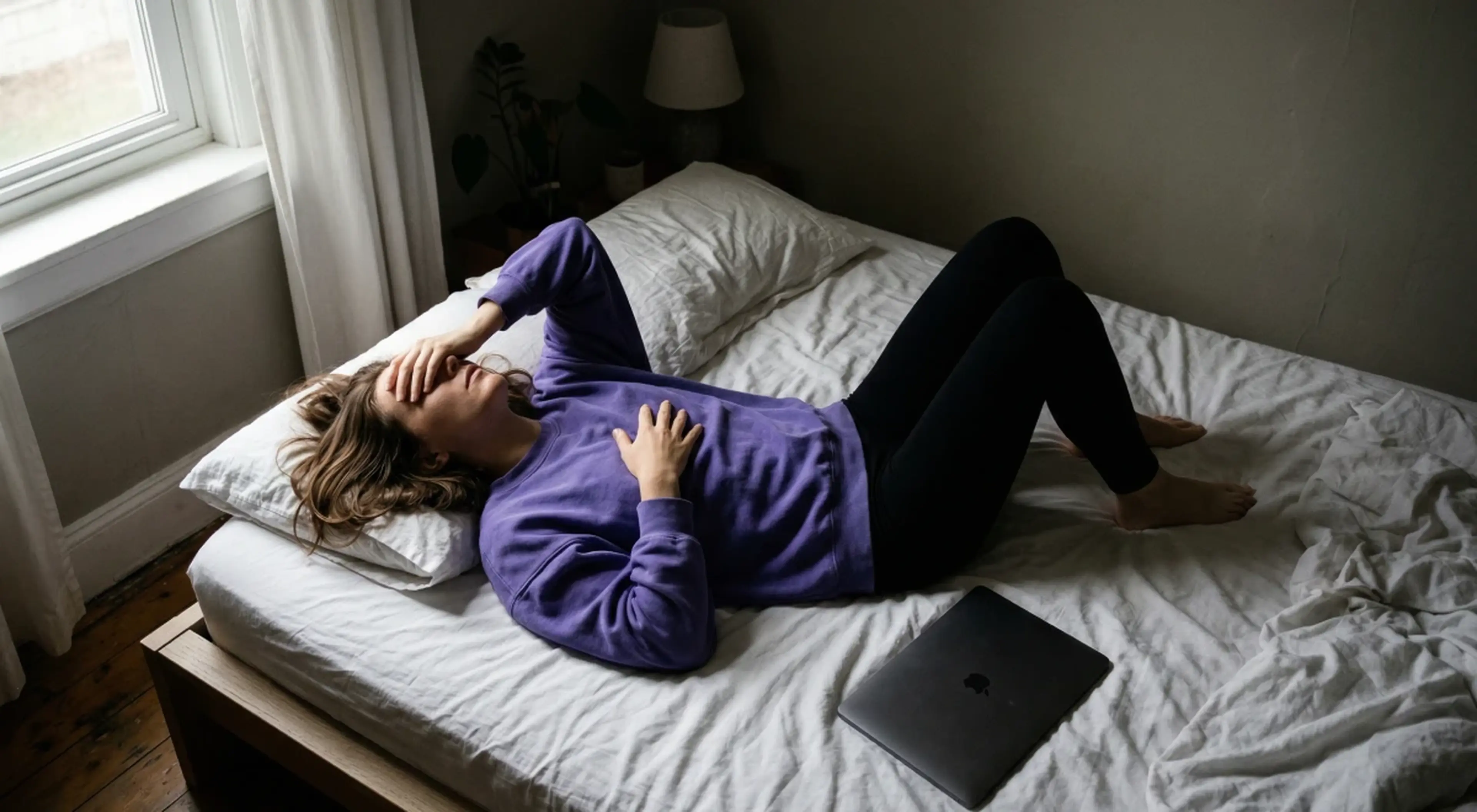 Woman in a purple hoodie lying on a rumpled bed holding her head in distress, laptop beside her, representing learned helplessness