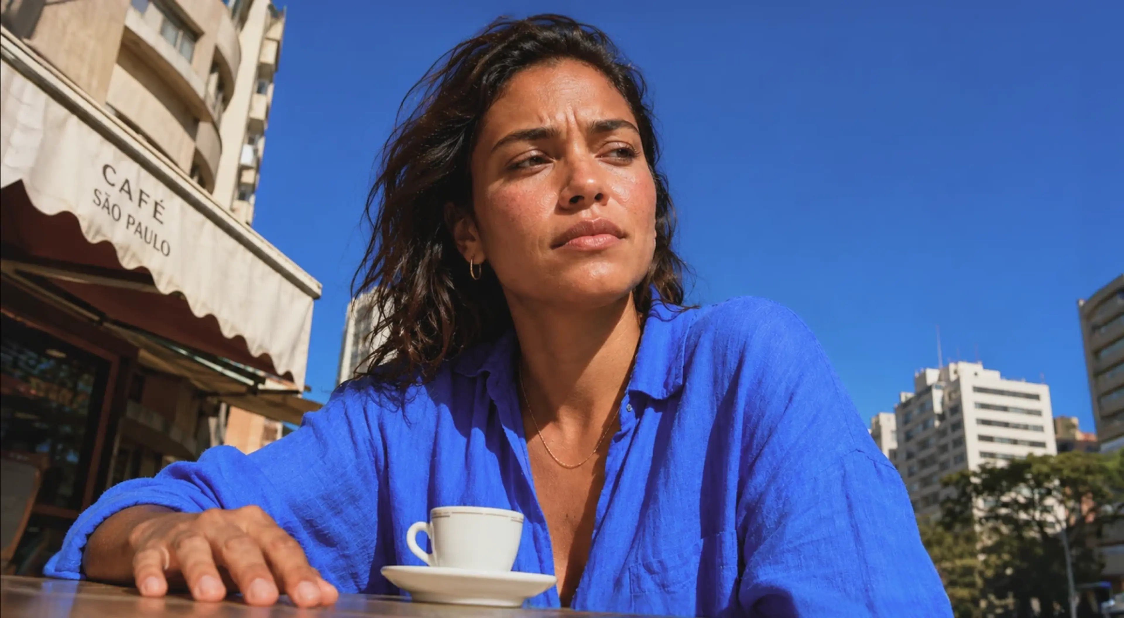 Woman in a blue shirt sitting alone at an outdoor café table with a coffee cup, looking away with a serious and uneasy expression under a clear blue sky