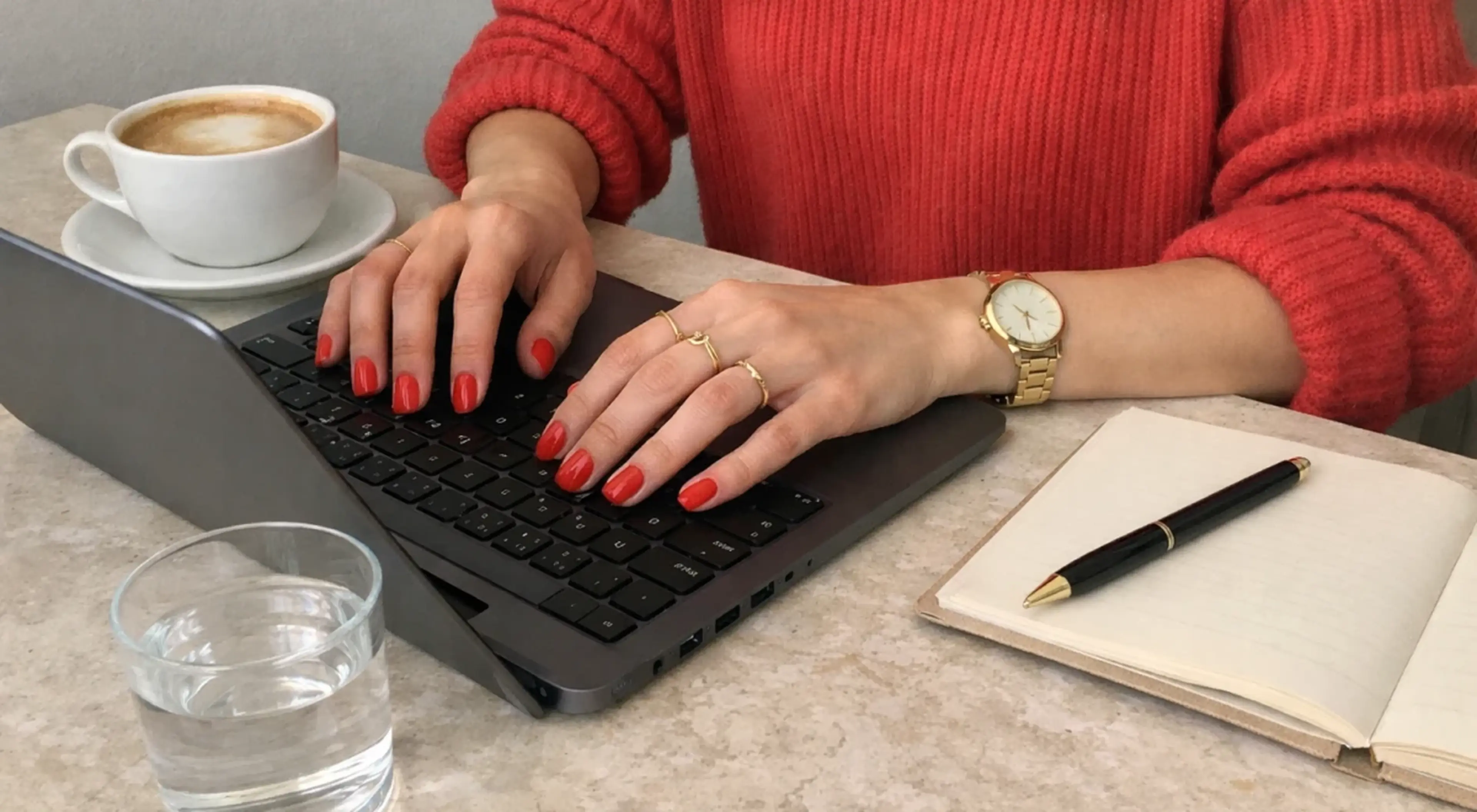 Successful woman in red sweater typing on laptop at marble desk with coffee, water glass, and open notebook as part of her morning routine