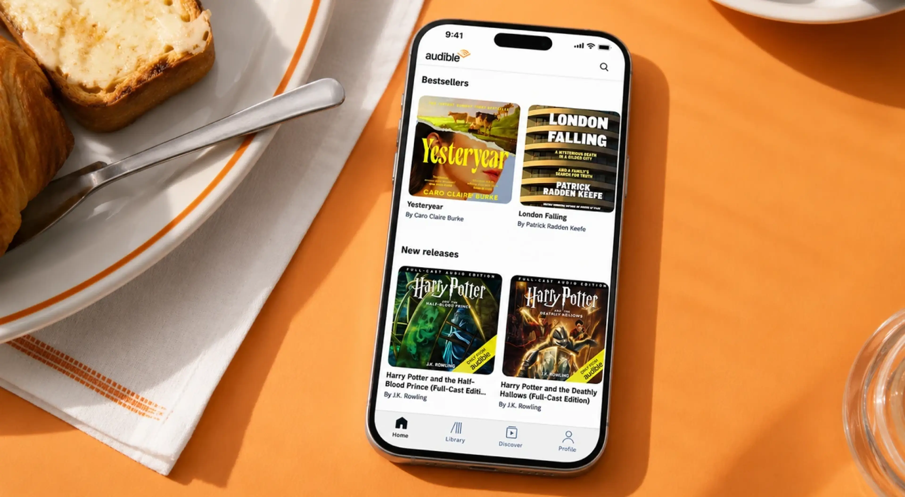Smartphone displaying the Audible app with bestseller and new release audiobook covers on an orange breakfast table surface with toast and a glass nearby