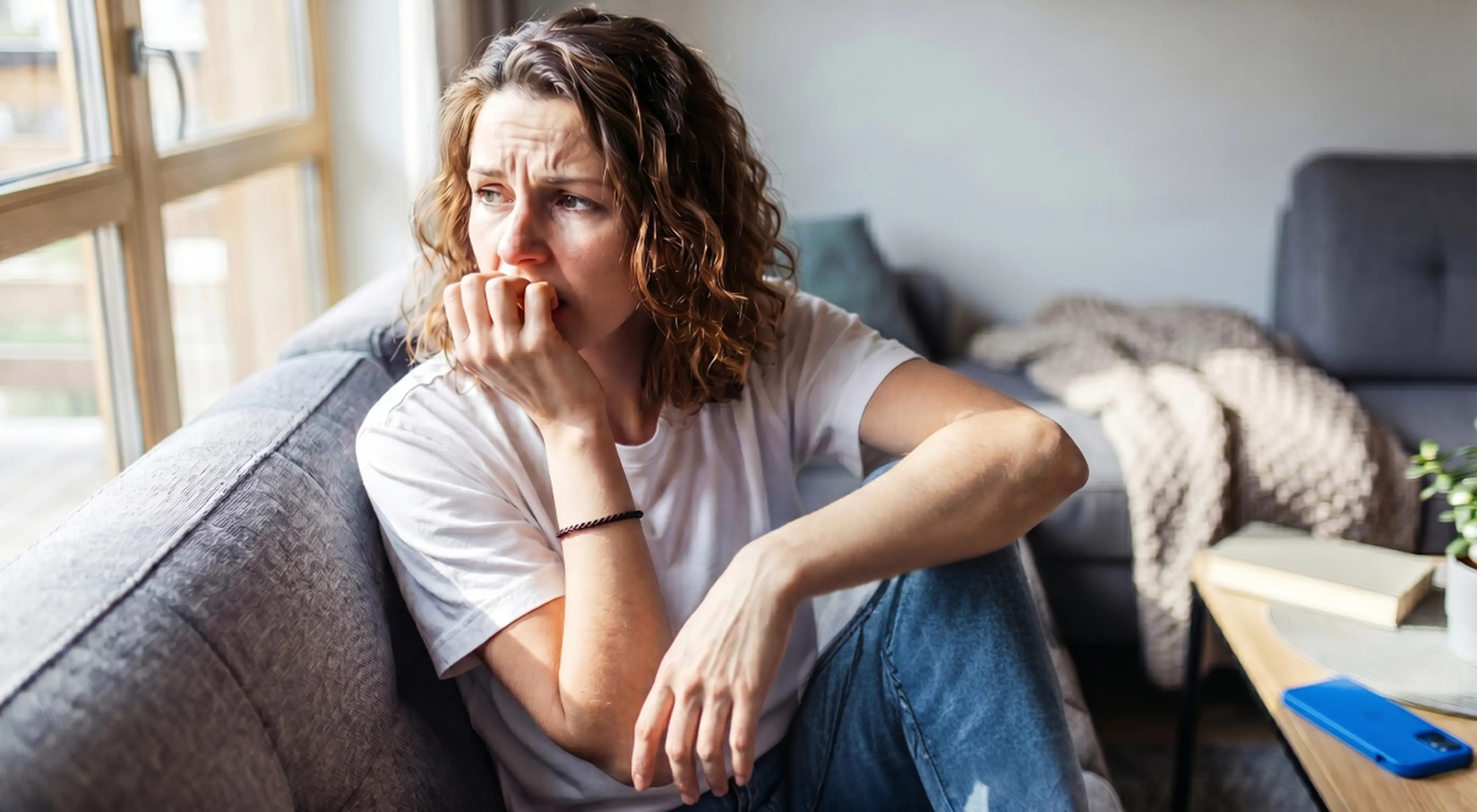 Worried woman with curly hair feeling anxious while sitting on the floor by a grey sofa, biting her nails and staring out the window in a bright living room