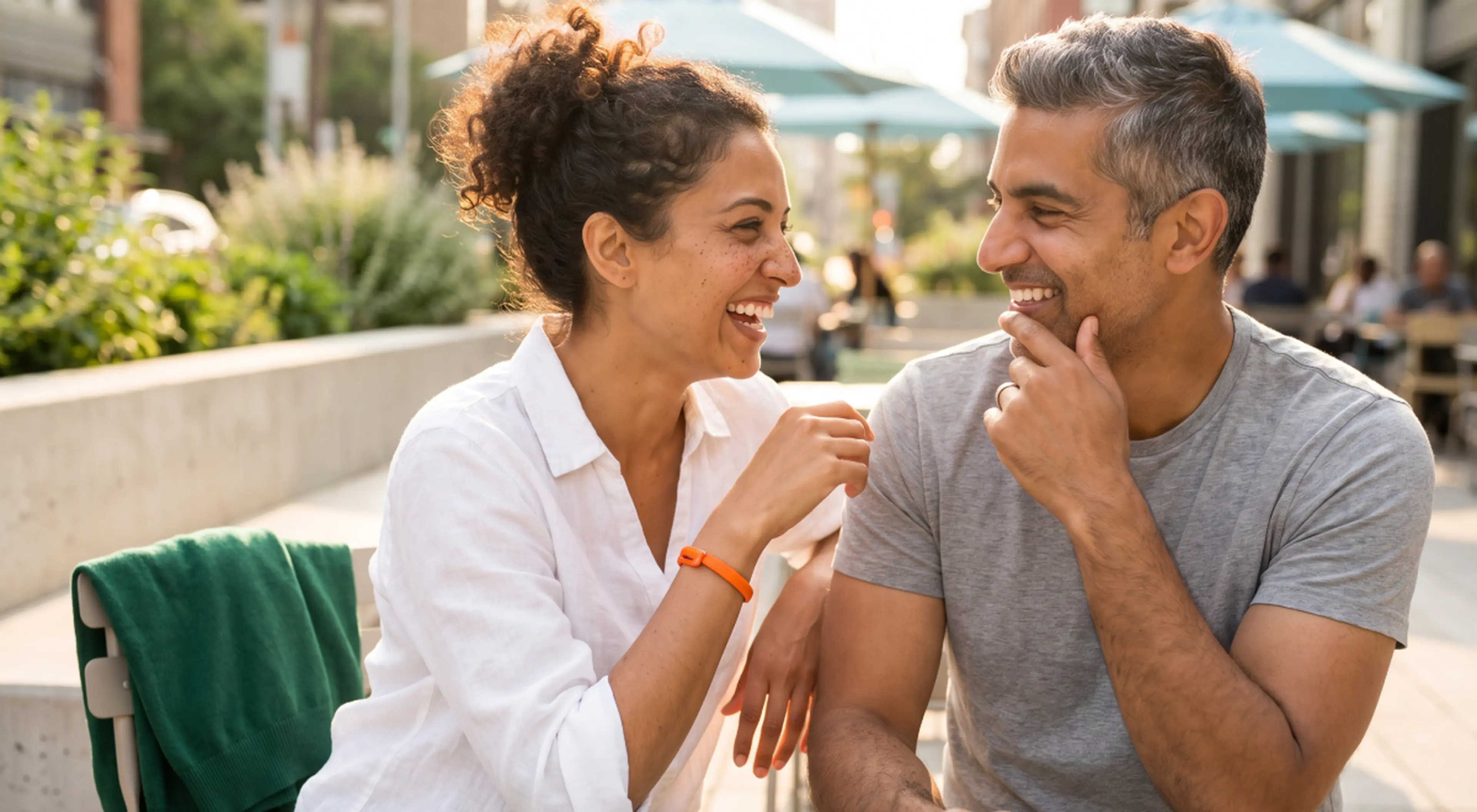 Smiling couple flirting and laughing together at an outdoor city café, the woman in a white shirt and the man in a grey t-shirt gazing at each other in warm sunlight