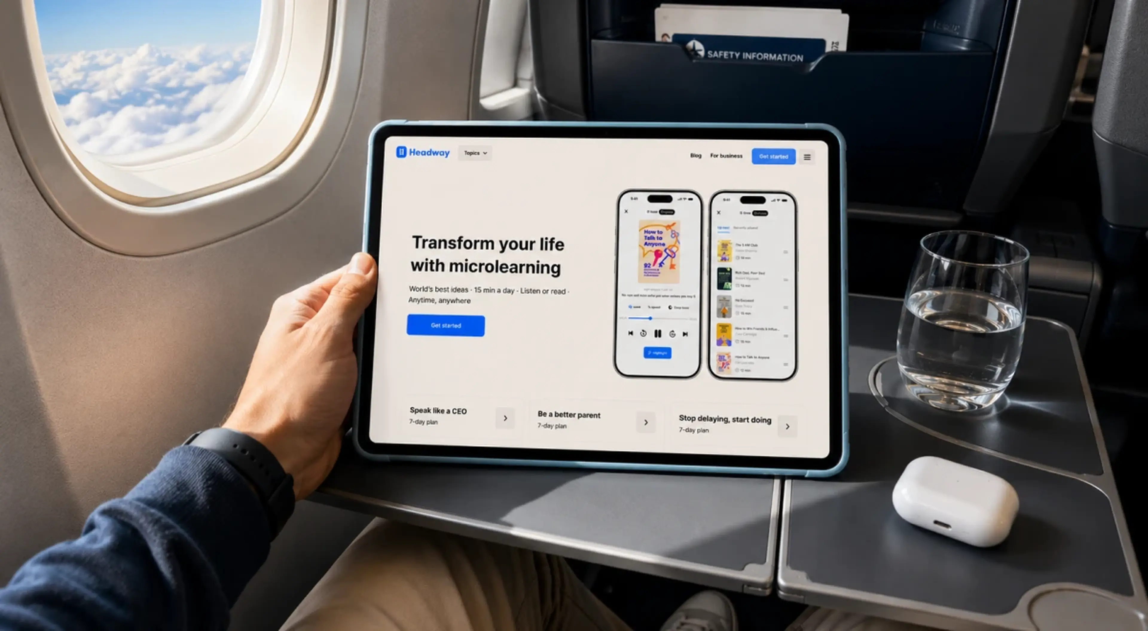 _Person holding a tablet displaying the Headway microlearning app on an airplane tray table with earbuds and a glass of water, clouds visible through the window