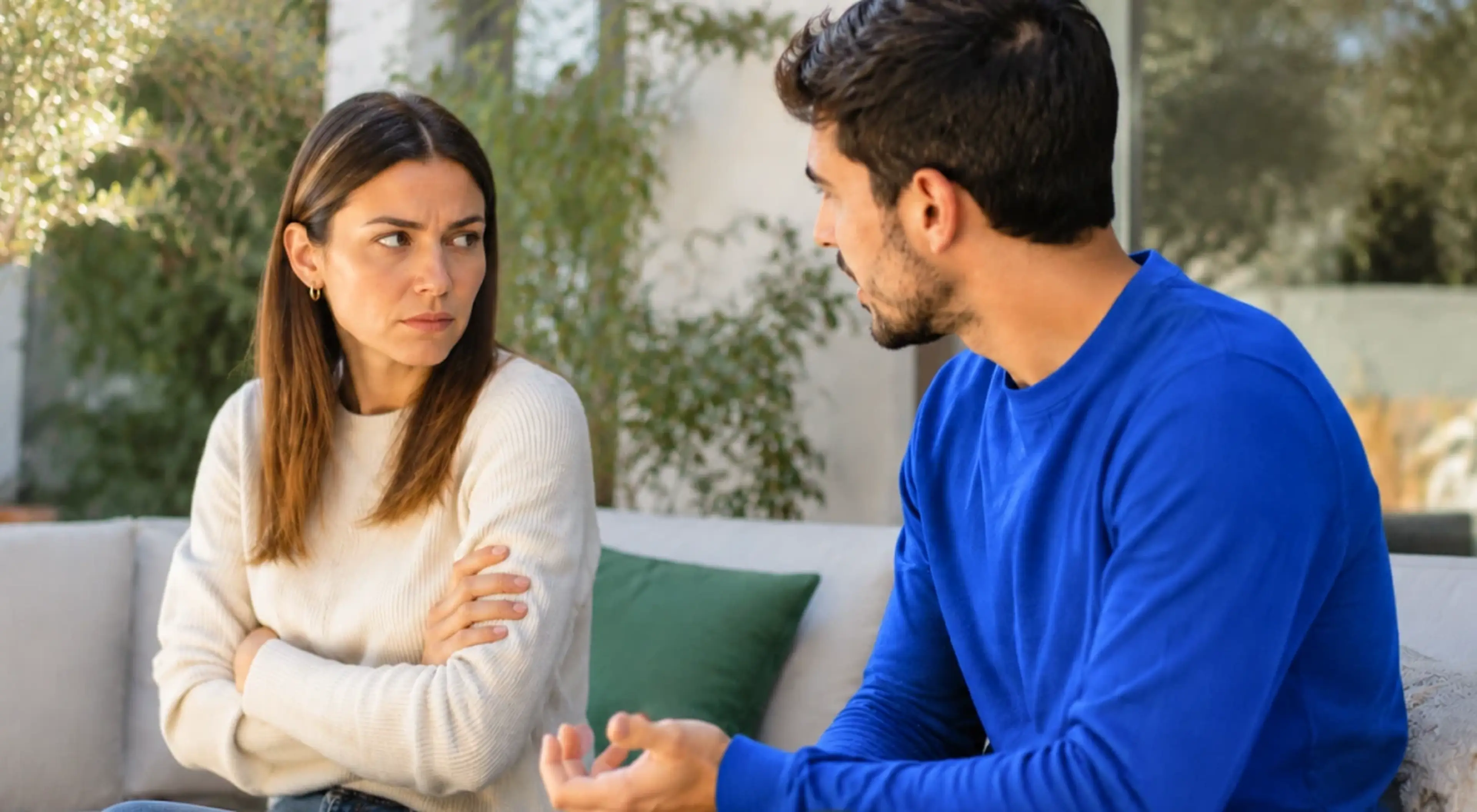 Woman with crossed arms looking away while a man in a blue sweater tries to talk to her on a couch, illustrating anger and conflict in a relationship