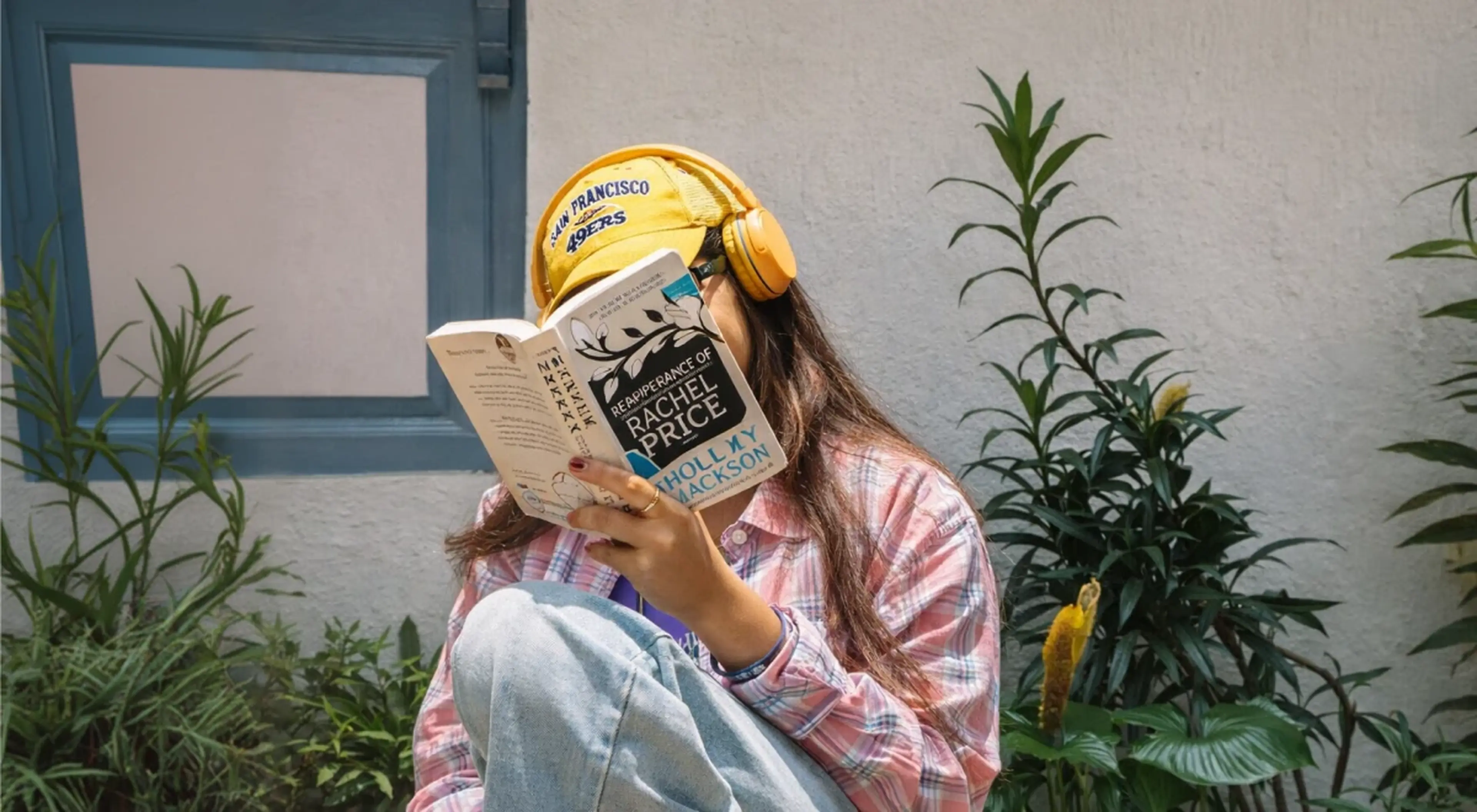Young woman in yellow headphones and pink plaid shirt listening and reading a book outdoors near green plants and a blue-framed window — thriller audiobooks