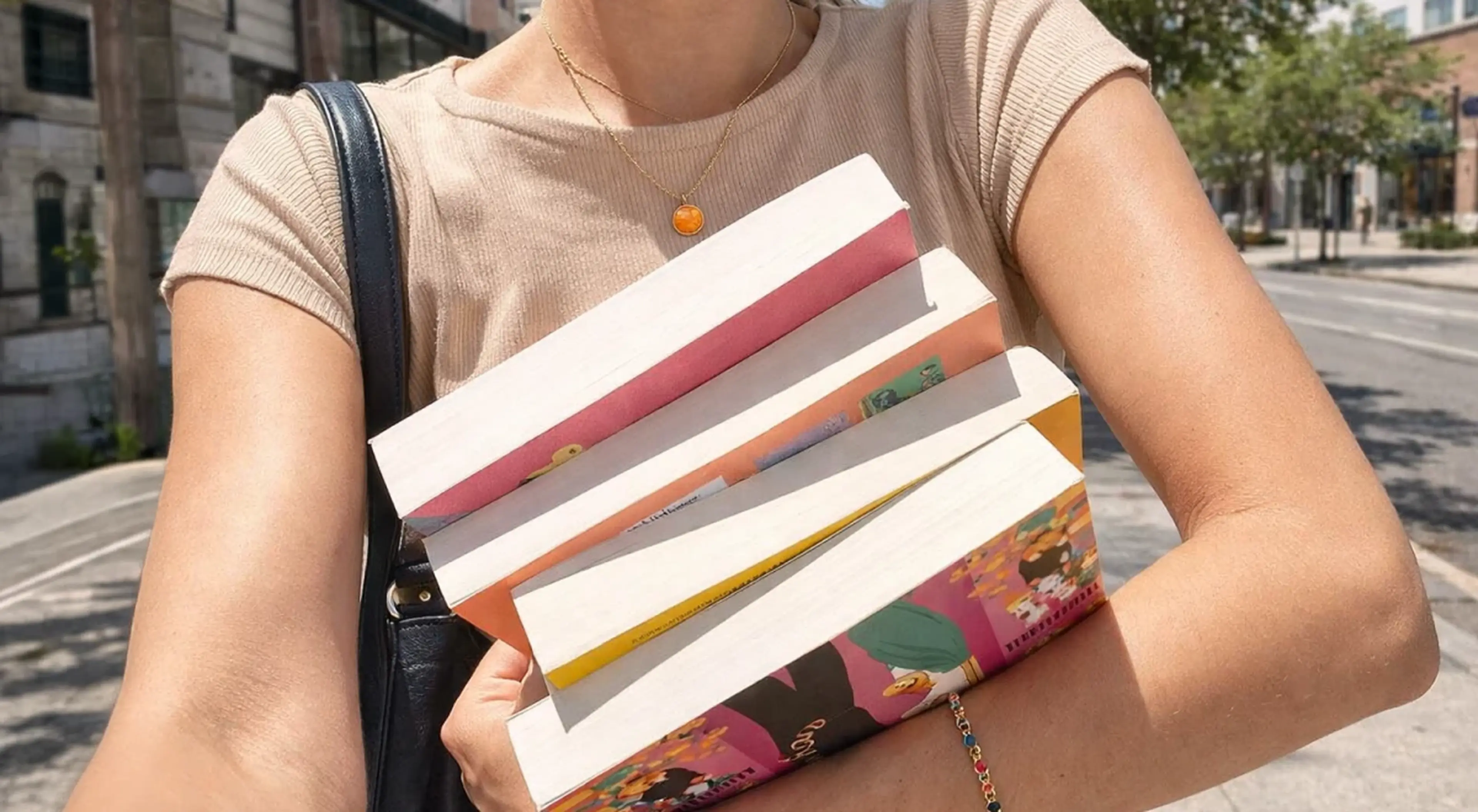 Woman in a beige top holding a stack of colorful books on a city street, wearing an orange pendant necklace — perfect visual for Rory Gilmore reading challenge recommendations