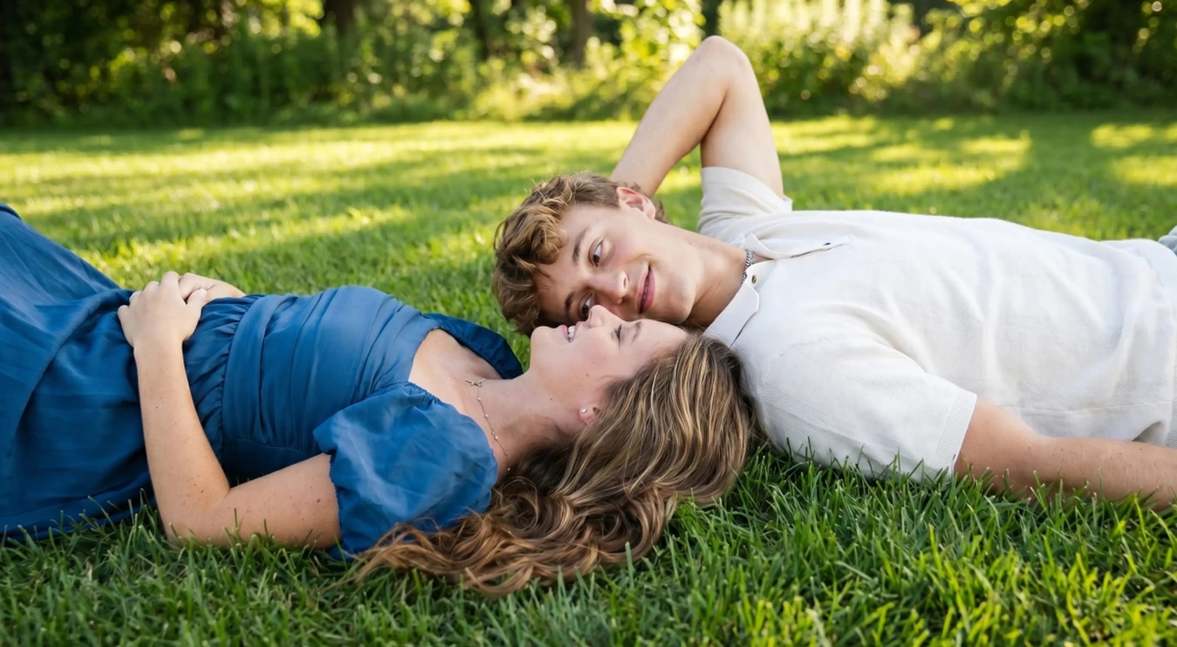 Young couple lying on green grass face to face in a romantic moment, woman in a blue dress and man in a white shirt, with lush green trees in the background