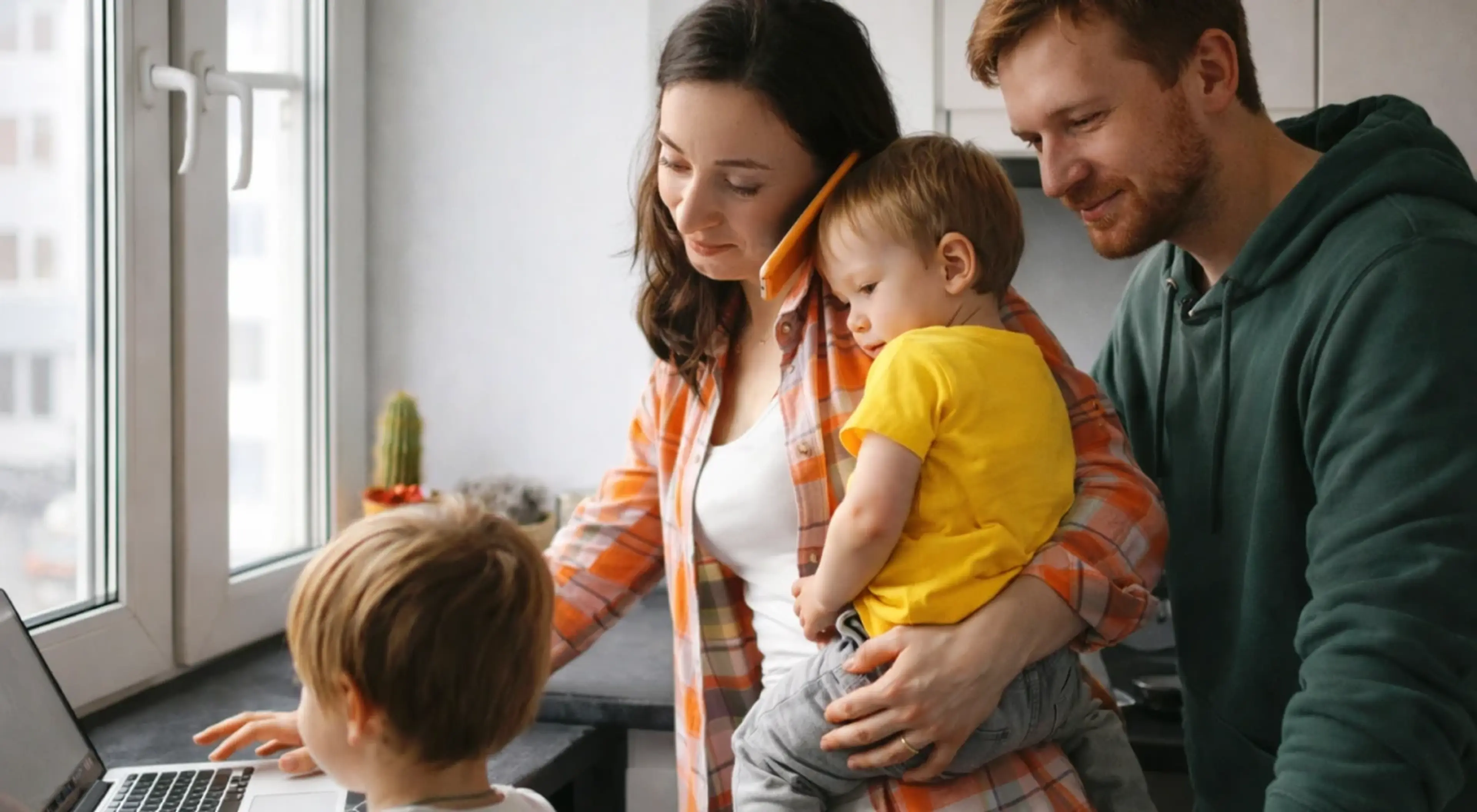 Mother in orange plaid shirt and father in green hoodie with toddler in yellow, child using laptop by window, best co-parenting app profile icon overlay