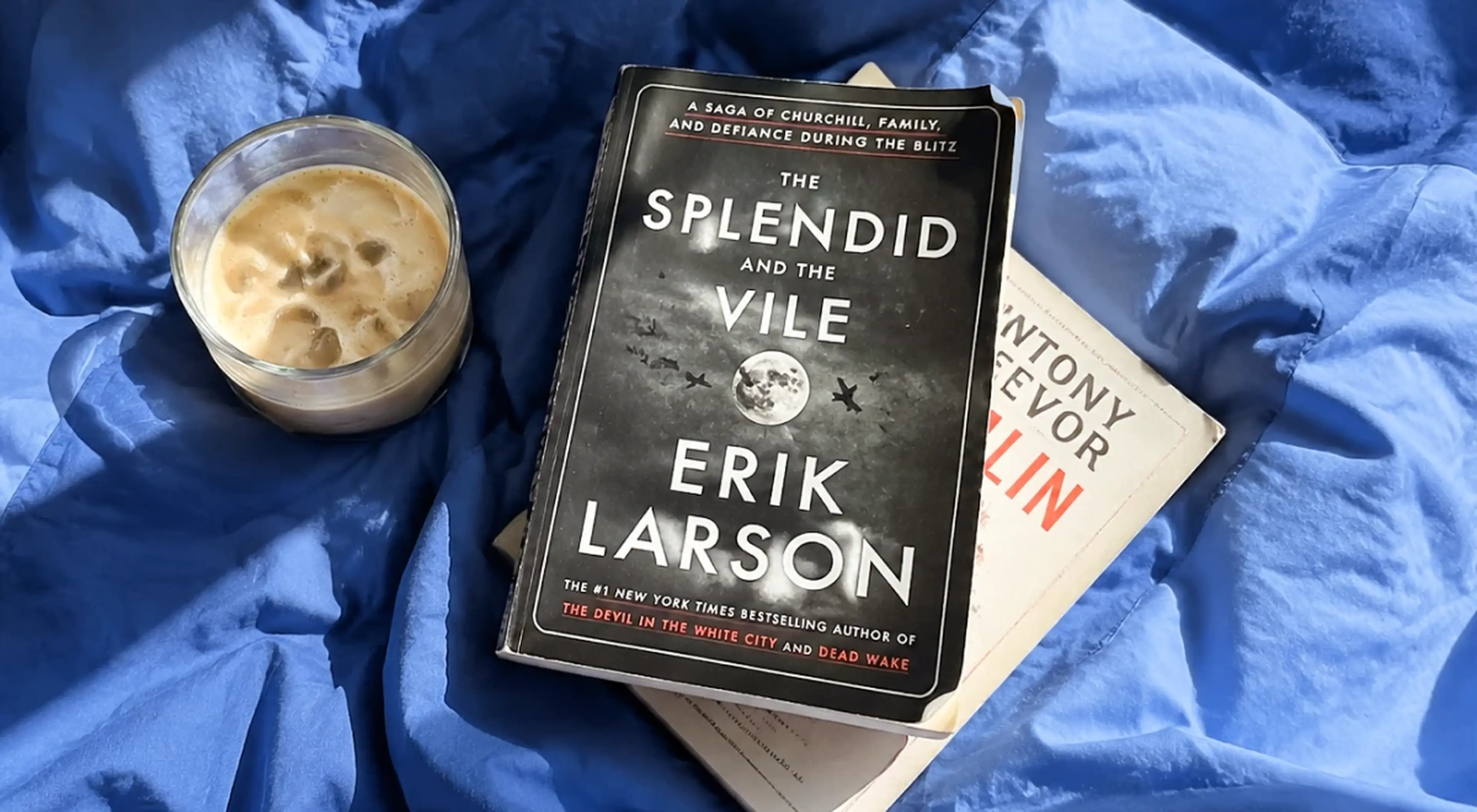 The Splendid and the Vile by Erik Larson placed on top of another book beside an iced coffee drink, all resting on blue fabric bedding (1)