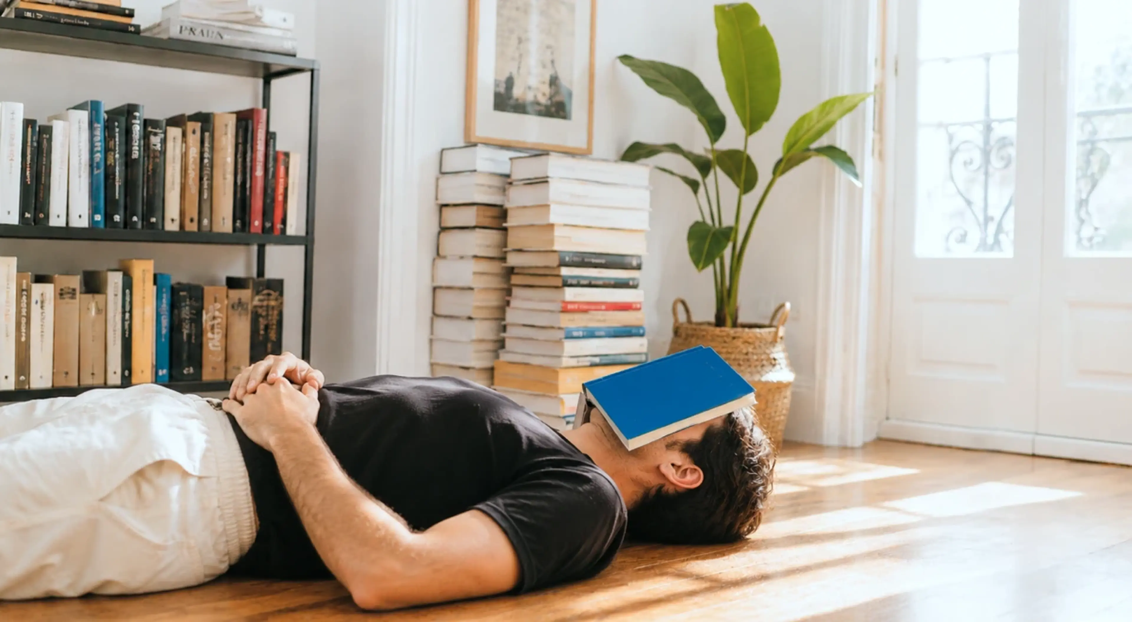 Man in a black shirt lying on the floor with a blue book on his face, surrounded by bookshelves and stacked books in a bright living room