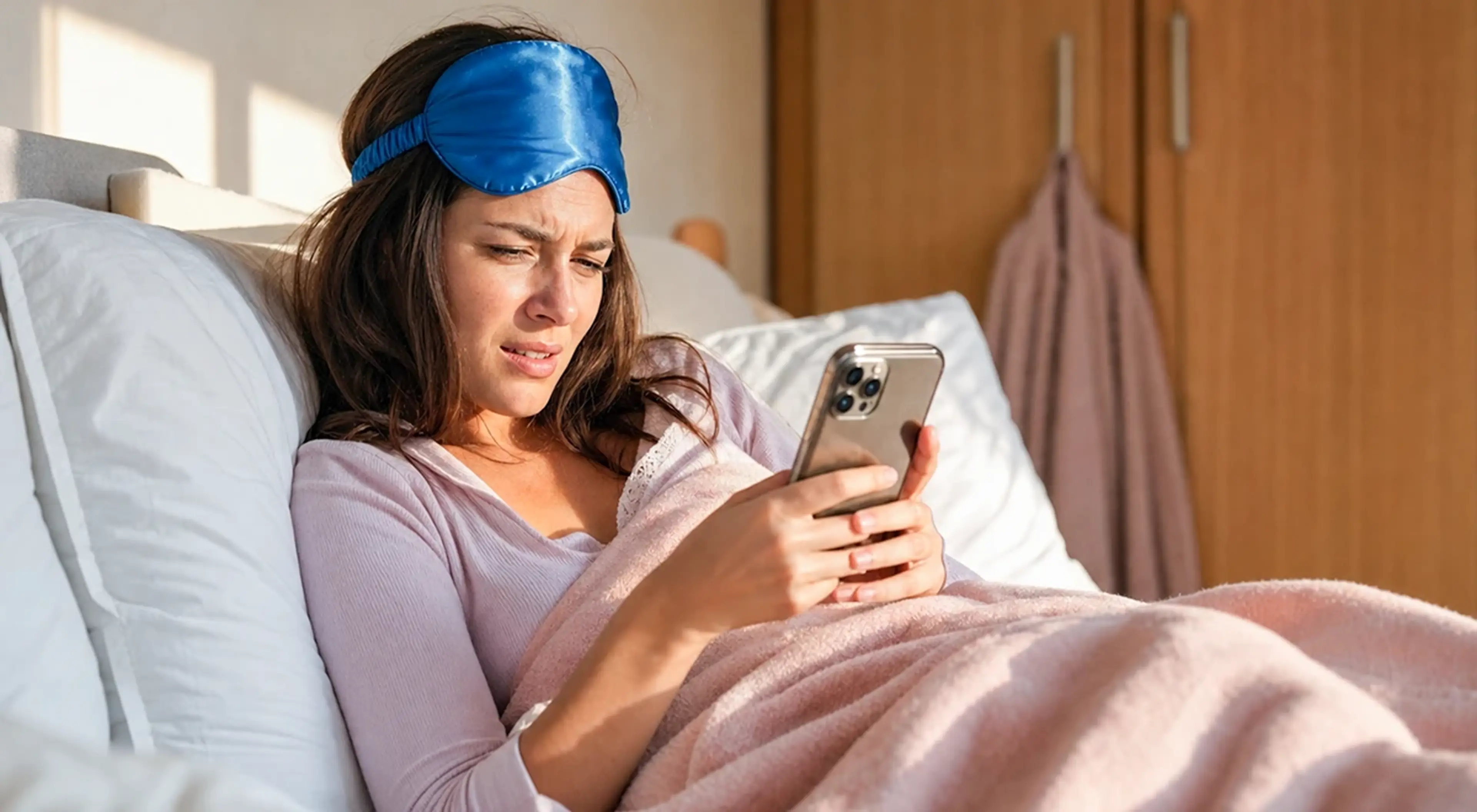 Woman lying in bed with a sleep mask on her forehead, looking upset while checking her phone, illustrating struggles to stop Instagram addiction at night