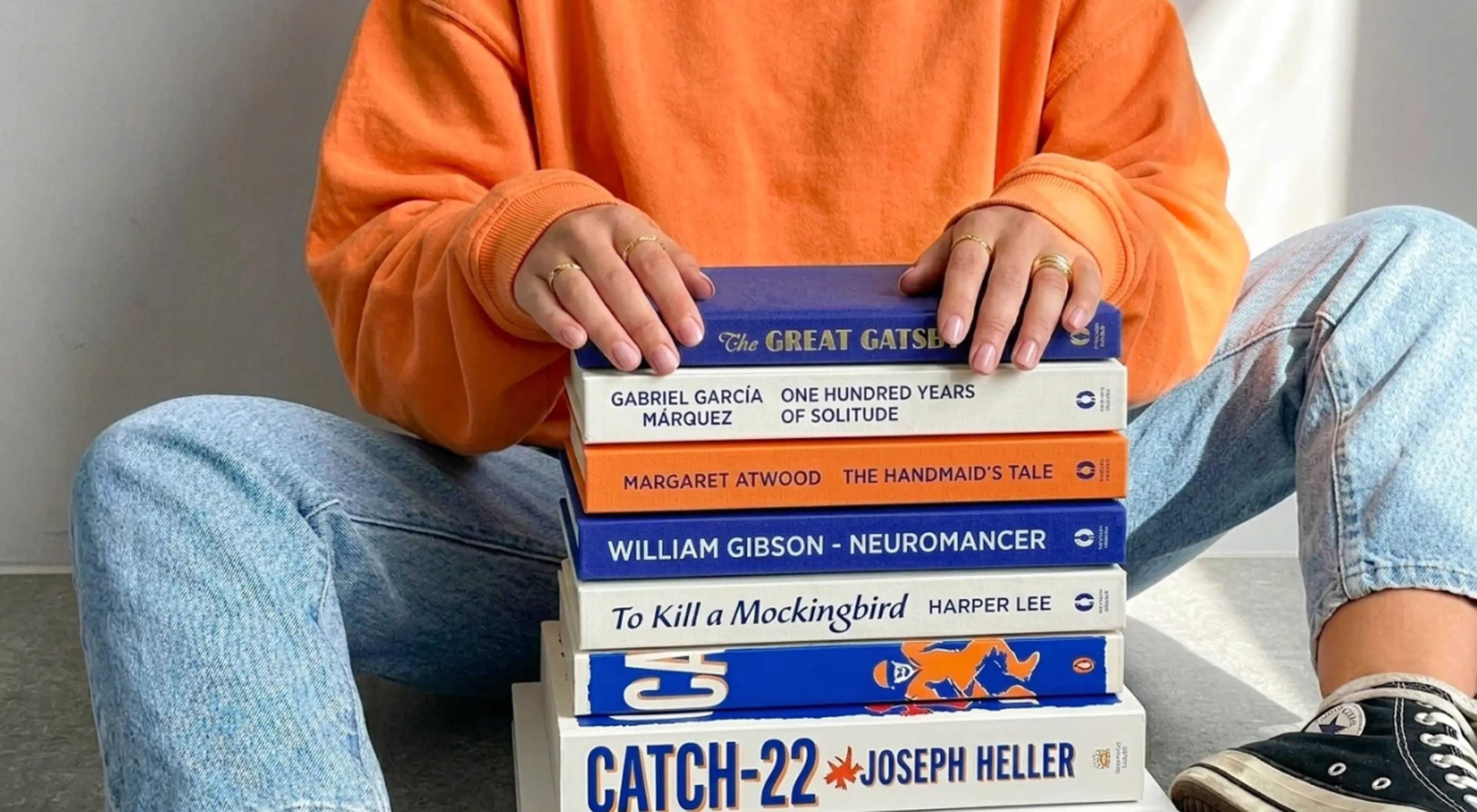 Person in an orange sweatshirt and jeans holding a stack of classic books including The Great Gatsby, Catch-22, To Kill a Mockingbird, and Neuromancer
