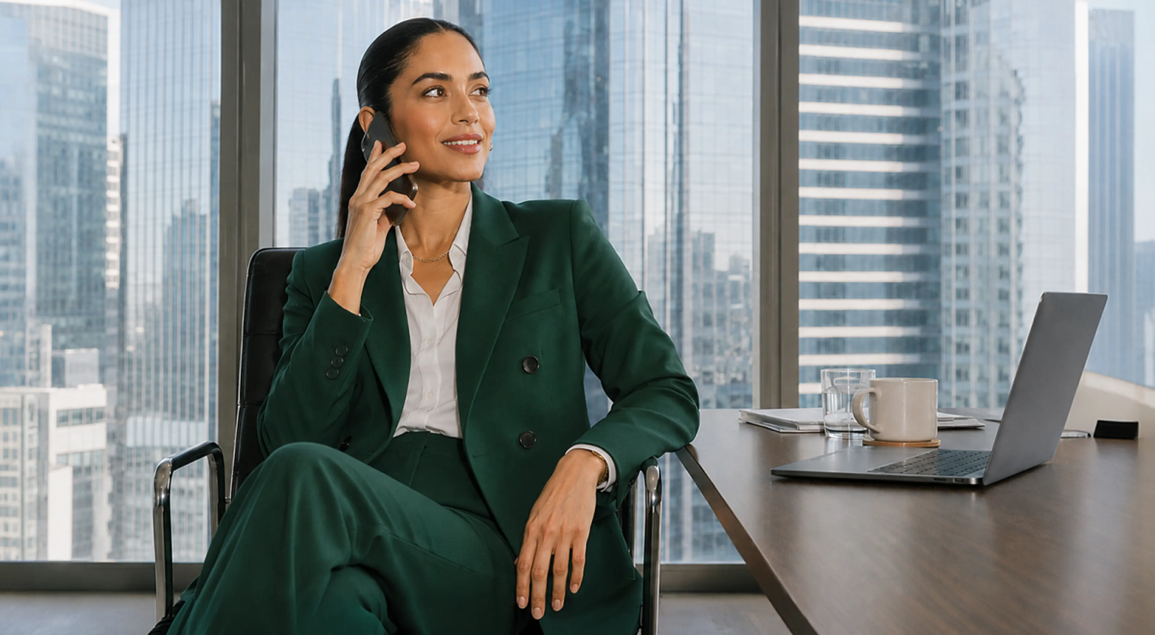 Confident businesswoman in a green suit talks on her phone in a high-rise office, ready to speak like a girlboss, with a laptop, coffee mug and city skyline behind her