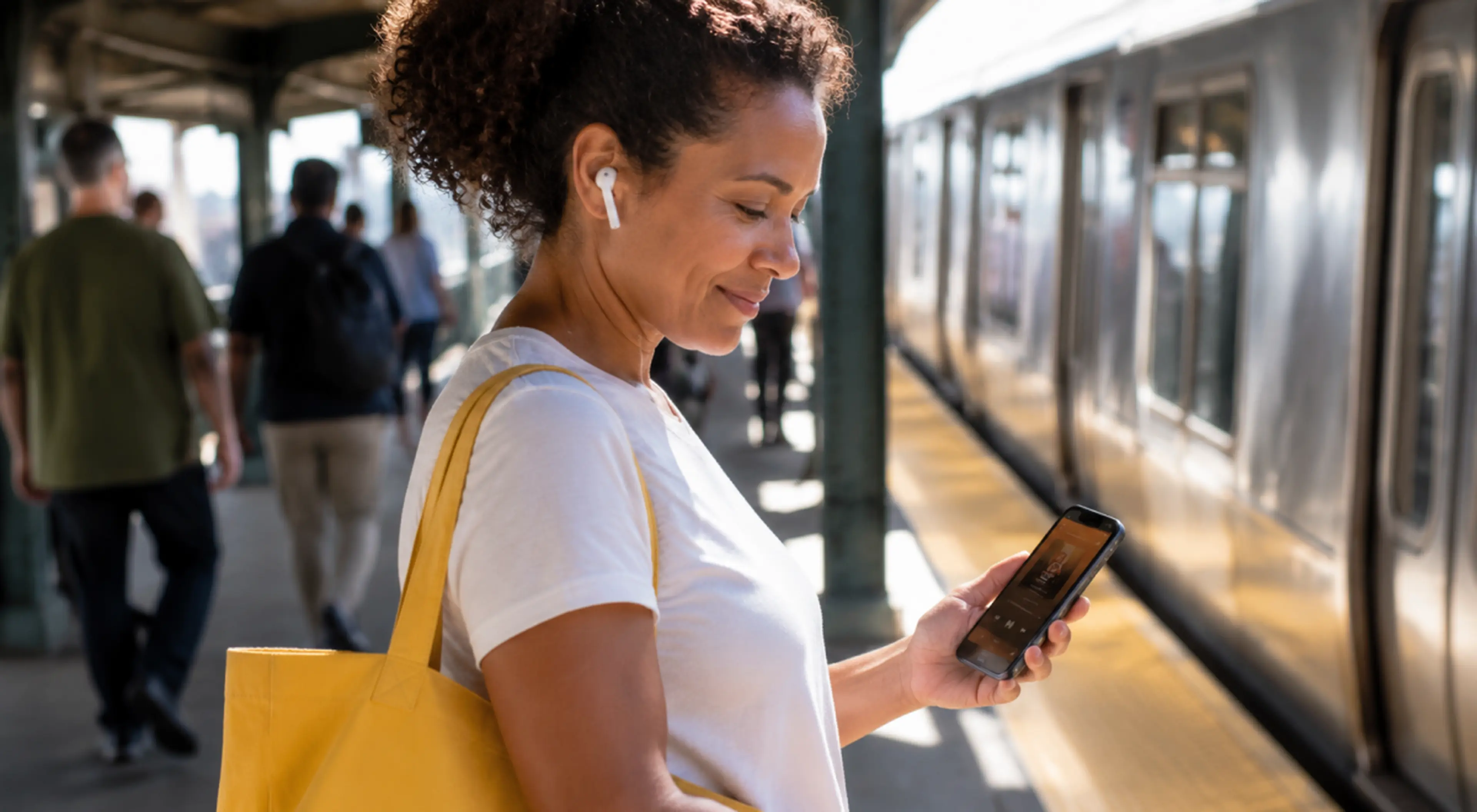 Woman with wireless earbuds and a yellow tote bag listening to podcasts on her phone while waiting on a sunny subway platform as a train and commuters pass by