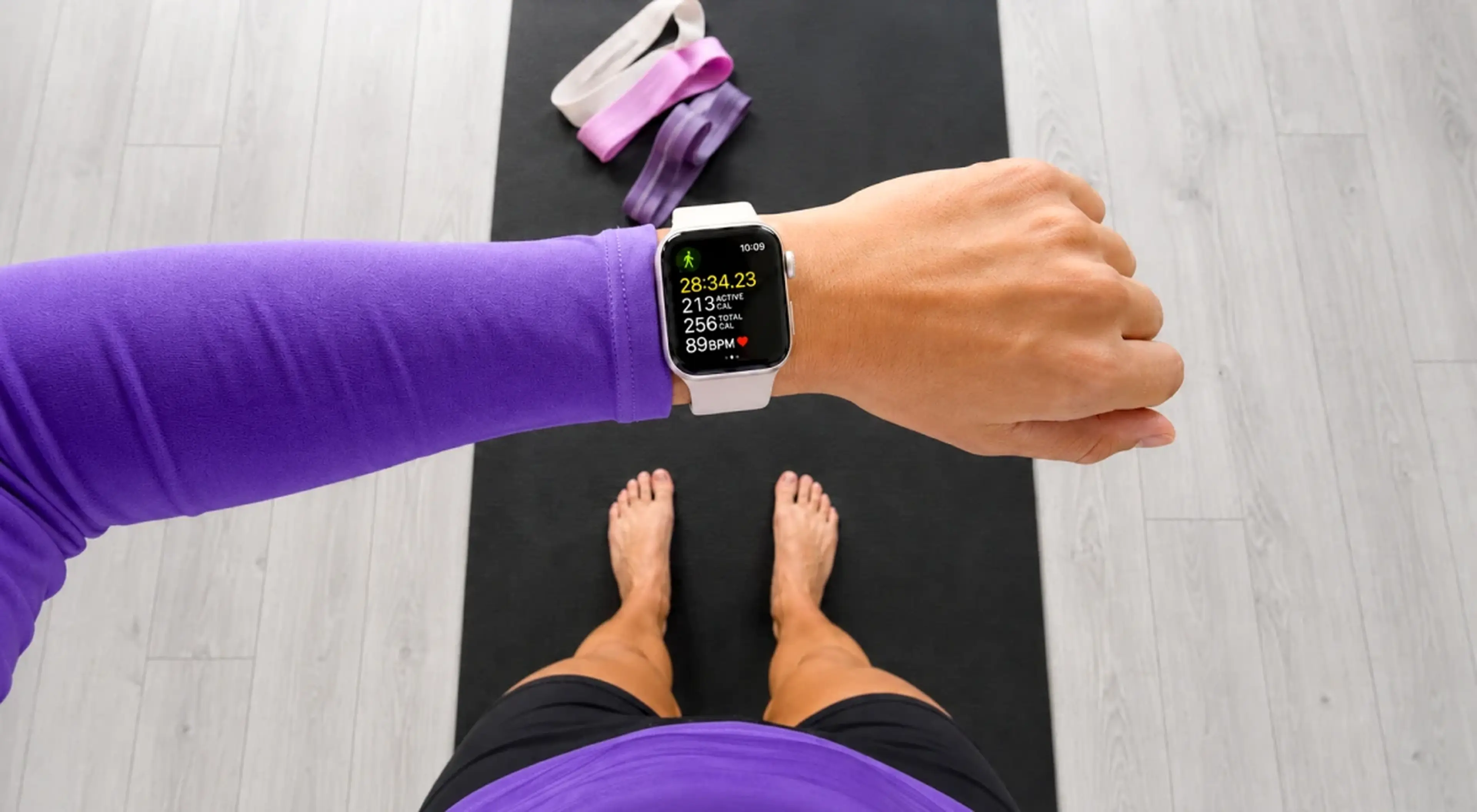Top-down view of a woman in purple activewear checking Apple Watch workout stats while standing on a black yoga mat with a resistance band, representing fitness discipline and tracking