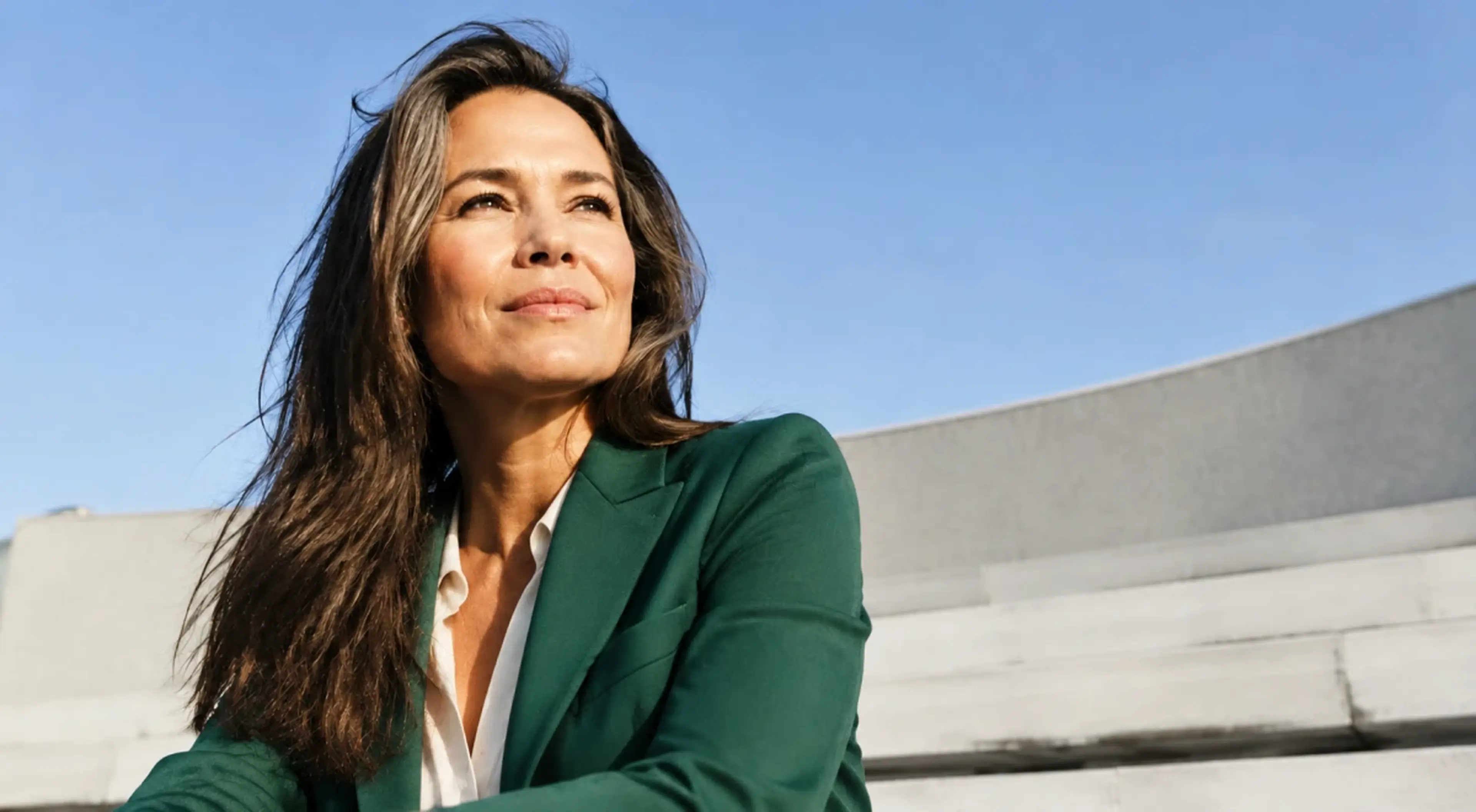 Confident woman in a green blazer looking up thoughtfully on a rooftop under a clear blue sky, conveying motivation and self-assurance