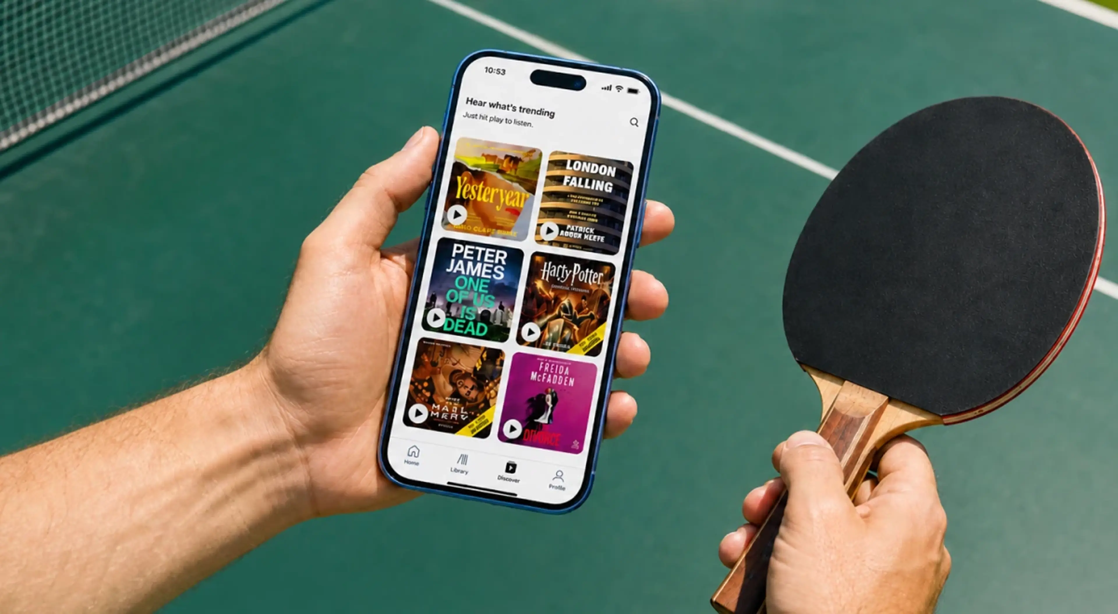 Man holding a smartphone displaying an audiobook app with trending titles and a ping-pong paddle, standing at a green table tennis table